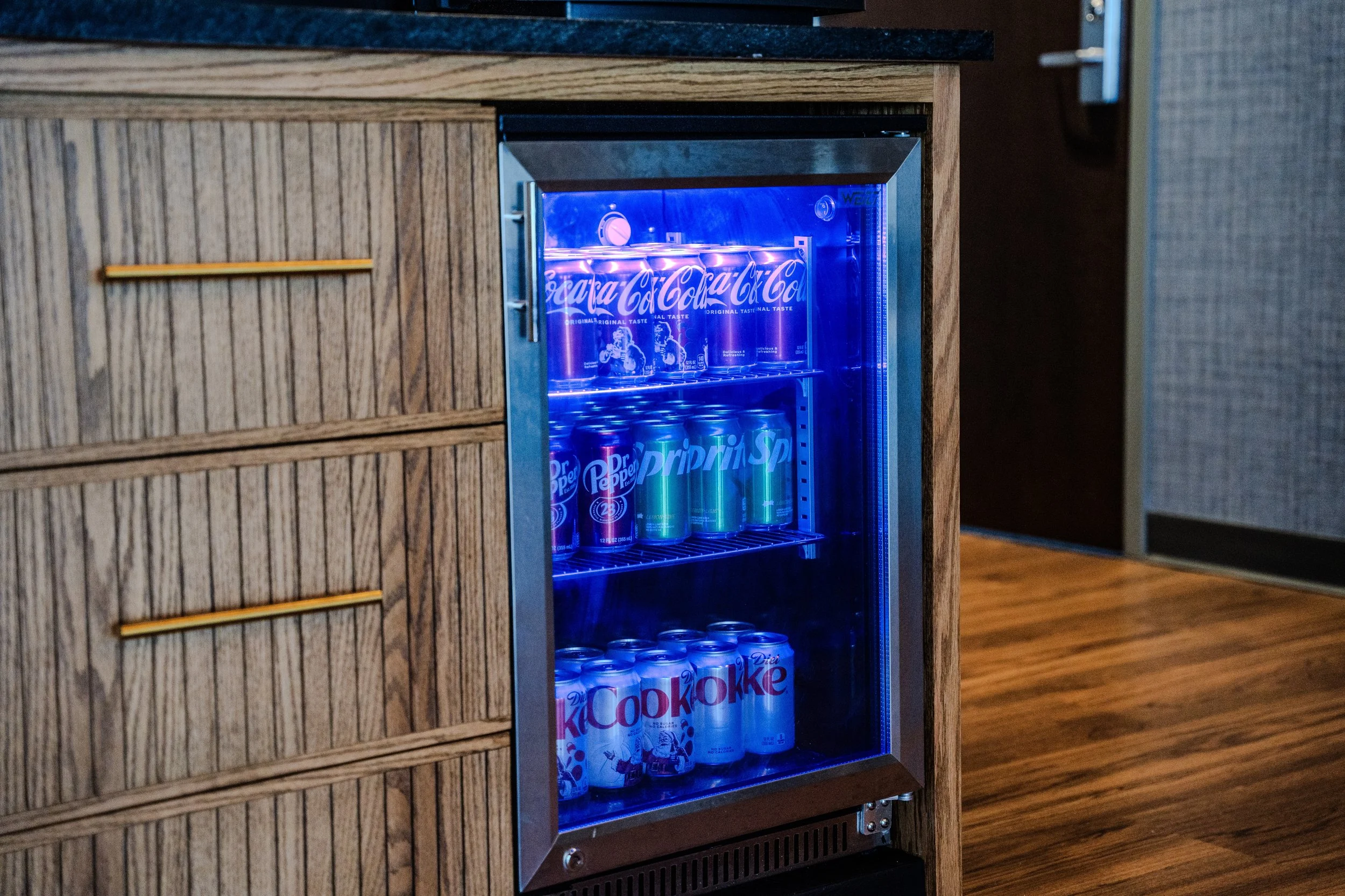A small refrigerator with a glass door illuminated by blue light, containing cans of Dr. Pepper and Coca-Cola, set in a room with wood flooring and a wooden cabinet nearby.