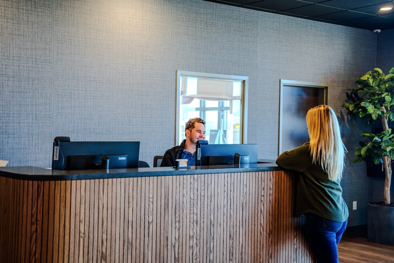 A woman with long blonde hair standing at a hotel reception desk, talking to a smiling male receptionist with short hair, in a modern lobby with wooden accents and a large potted plant.