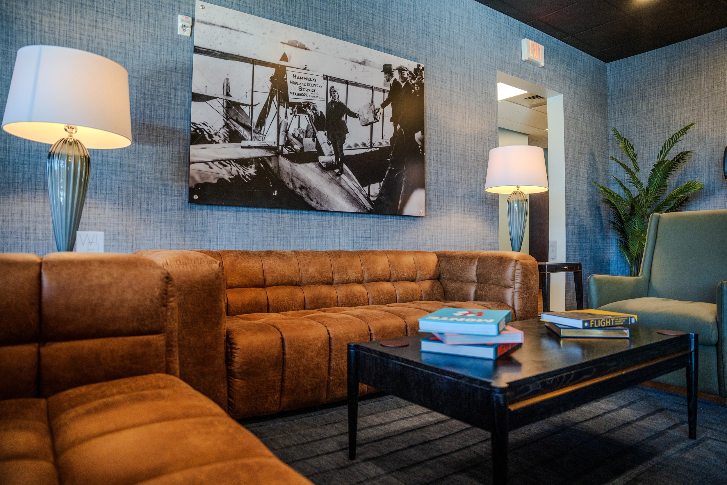 Living room with brown tufted leather sofa, black coffee table with books, blue walls, white lamps, large black and white photo of a historical scene on the wall, a green potted plant, and an exit door.