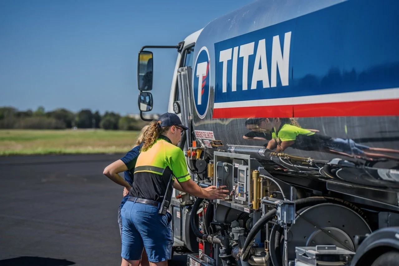 A worker in a bright yellow safety vest and cap operates machinery on a large Titan-branded tanker truck parked on a runway or tarmac with a clear blue sky and grassy landscape in the background.