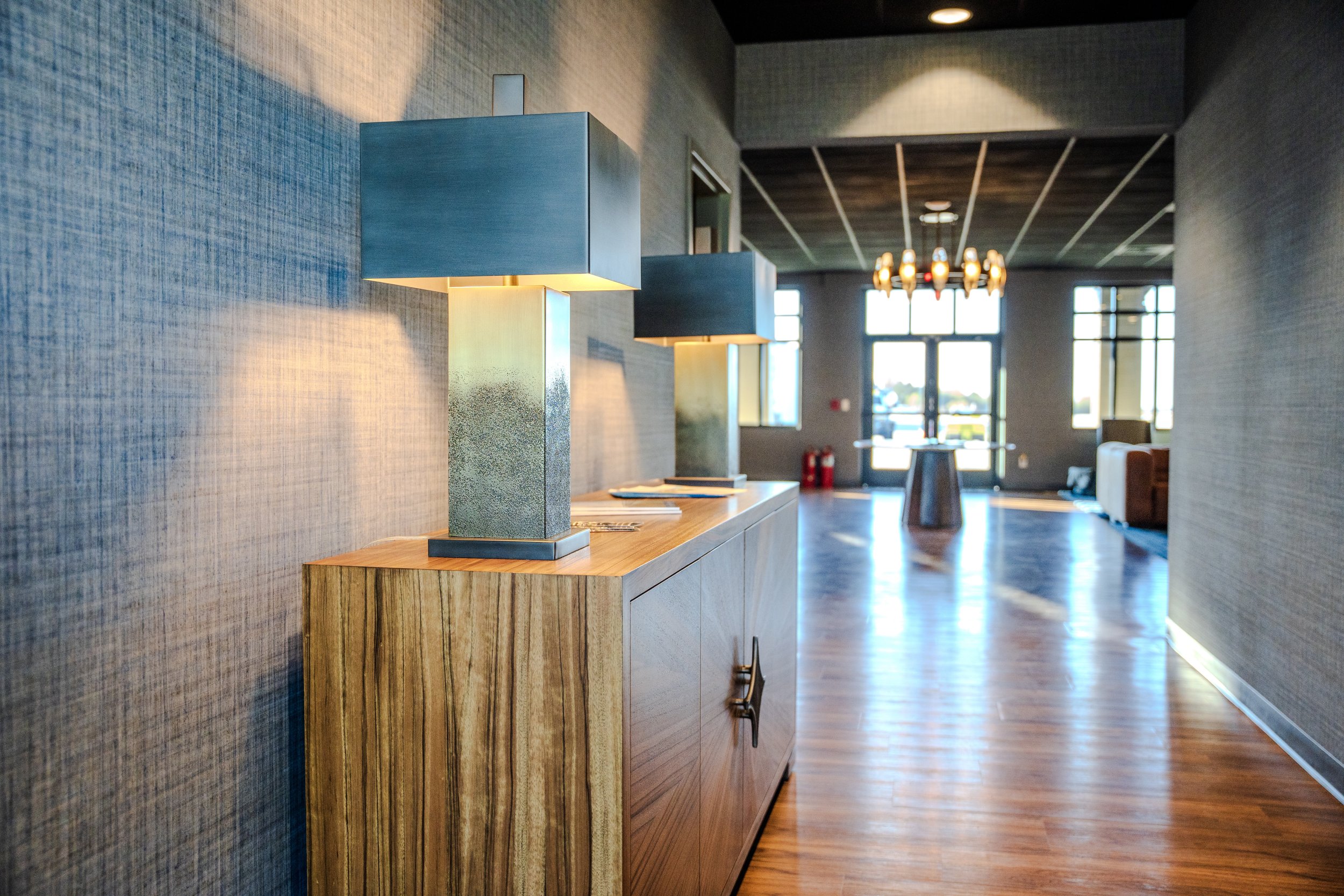 A modern, well-lit interior with wooden flooring, textured dark gray walls, and blue and gray lamps on a wooden sideboard. Large windows in the background let in natural light, and a dining area with a chandelier is visible.