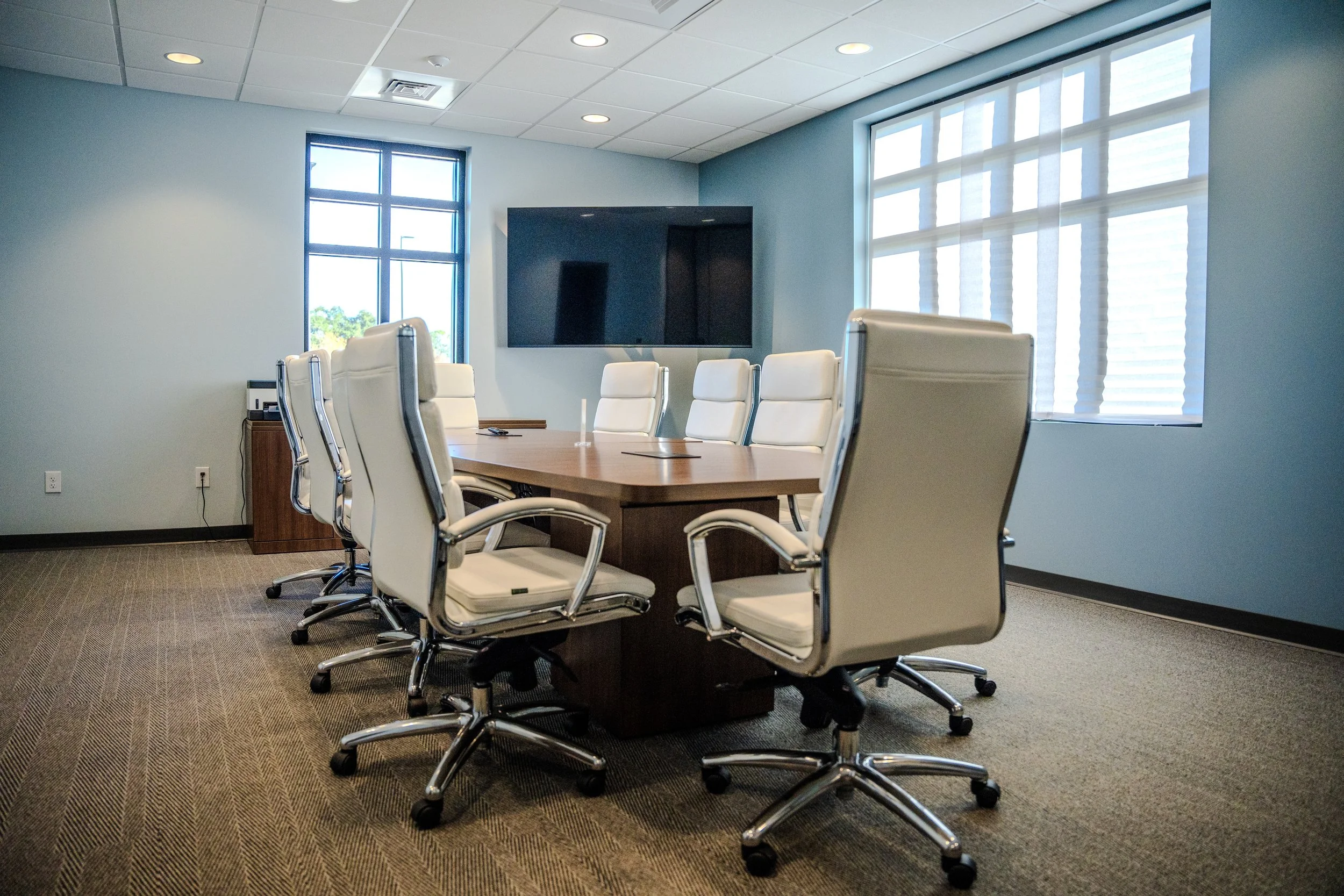 A modern conference room with a large wooden table surrounded by white leather office chairs, a wall-mounted television, and large windows with blinds overlooking trees outside.