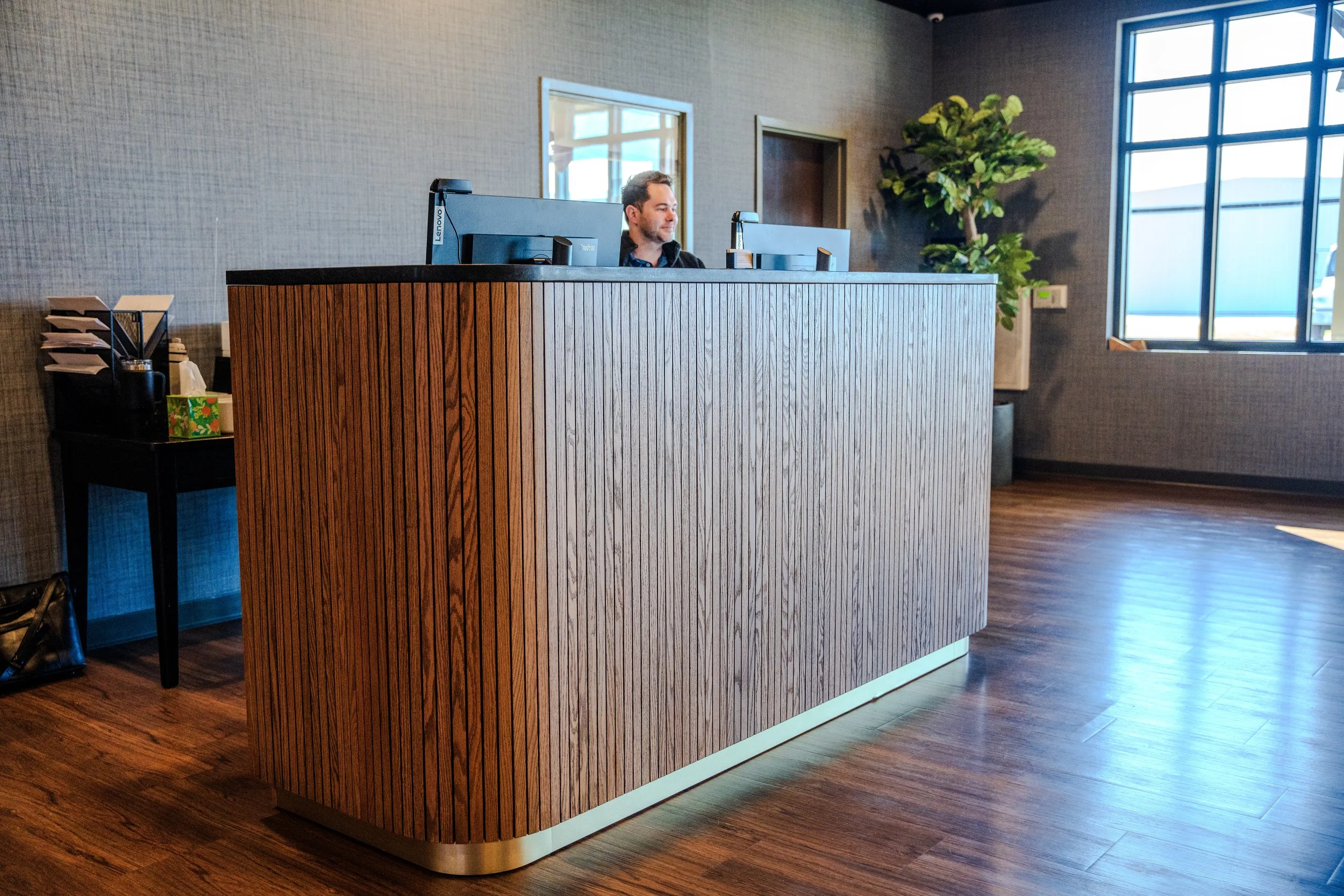 Reception desk with computer monitors, a potted plant in the corner, and a window letting in natural light.