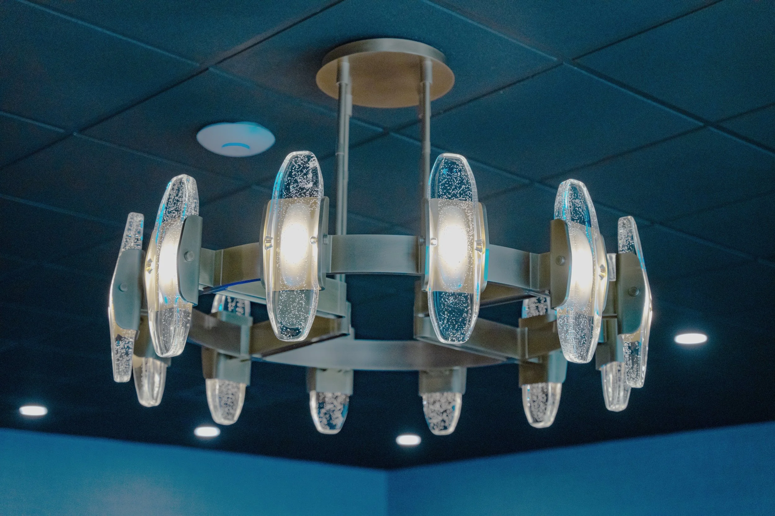 A modern chandelier with clear, elongated, oval-shaped glass pieces with bubbles inside, hanging from a round metallic fixture on a dark ceiling.