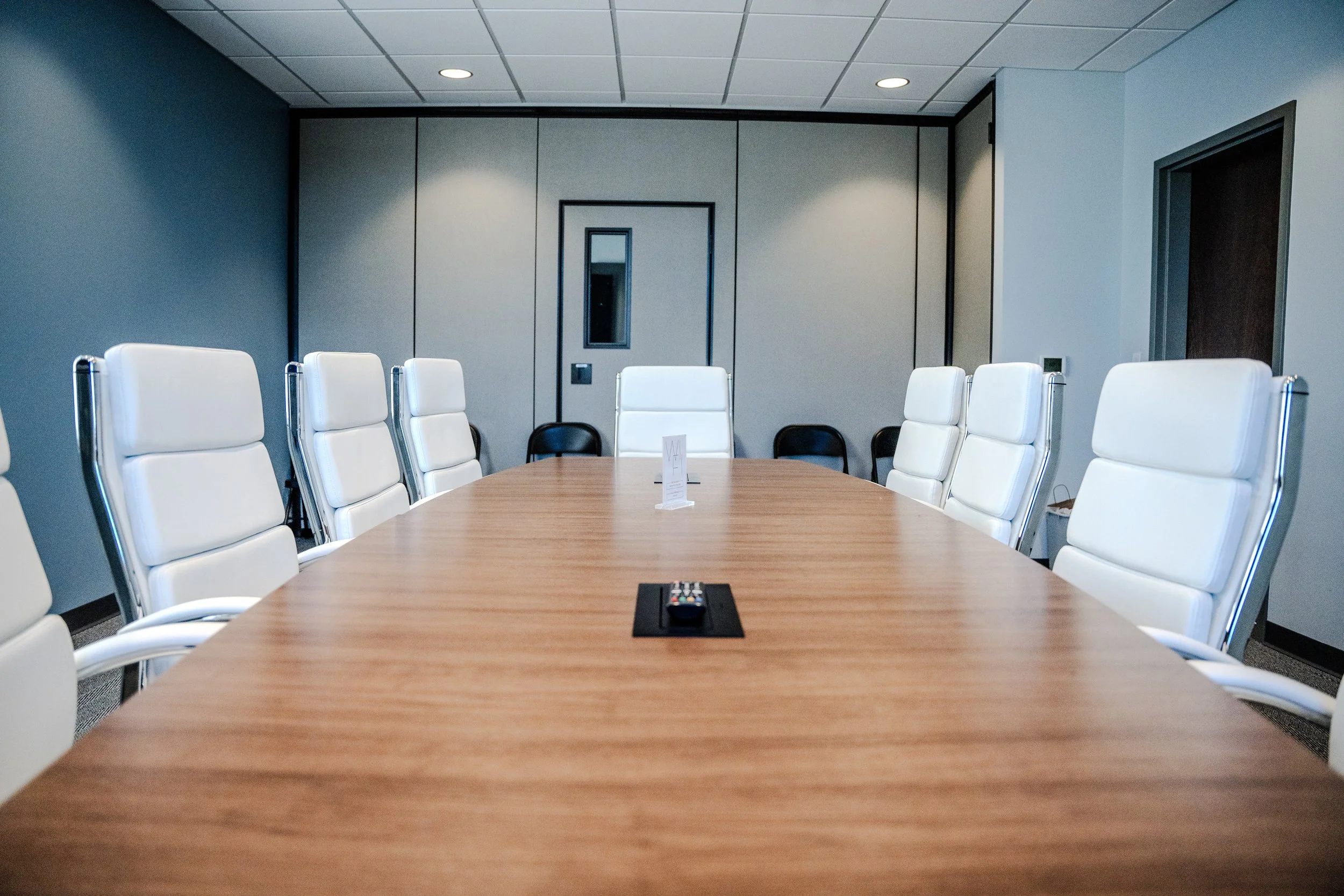 A conference room with a long wooden table surrounded by white leather chairs. There is a doorway with a mirror on the back wall of the room. A remote control is on the table.