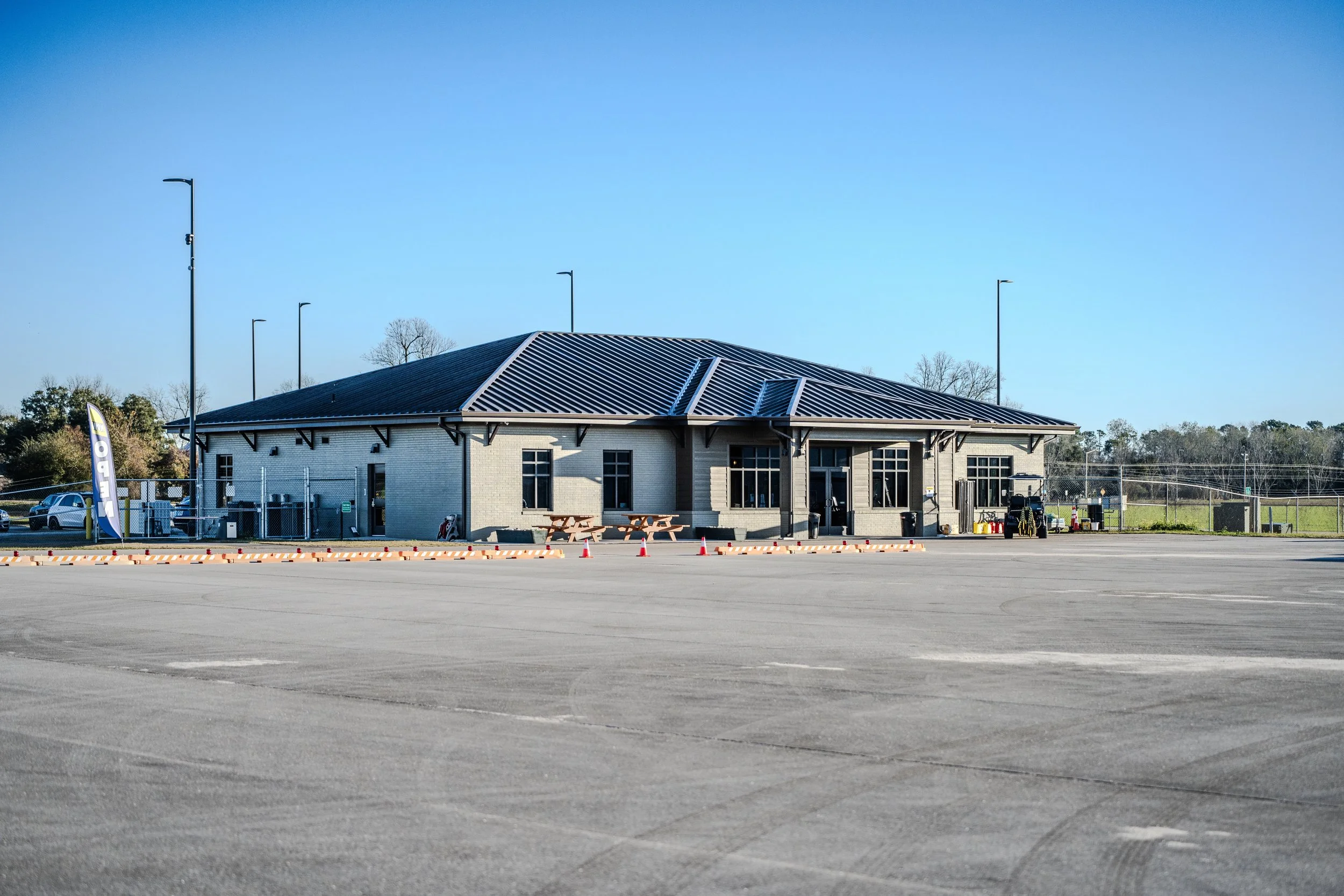 A single-story building with a dark metal roof, large windows, picnic tables outside, and a parking lot with orange traffic cones and a few cars. The sky is clear and blue.