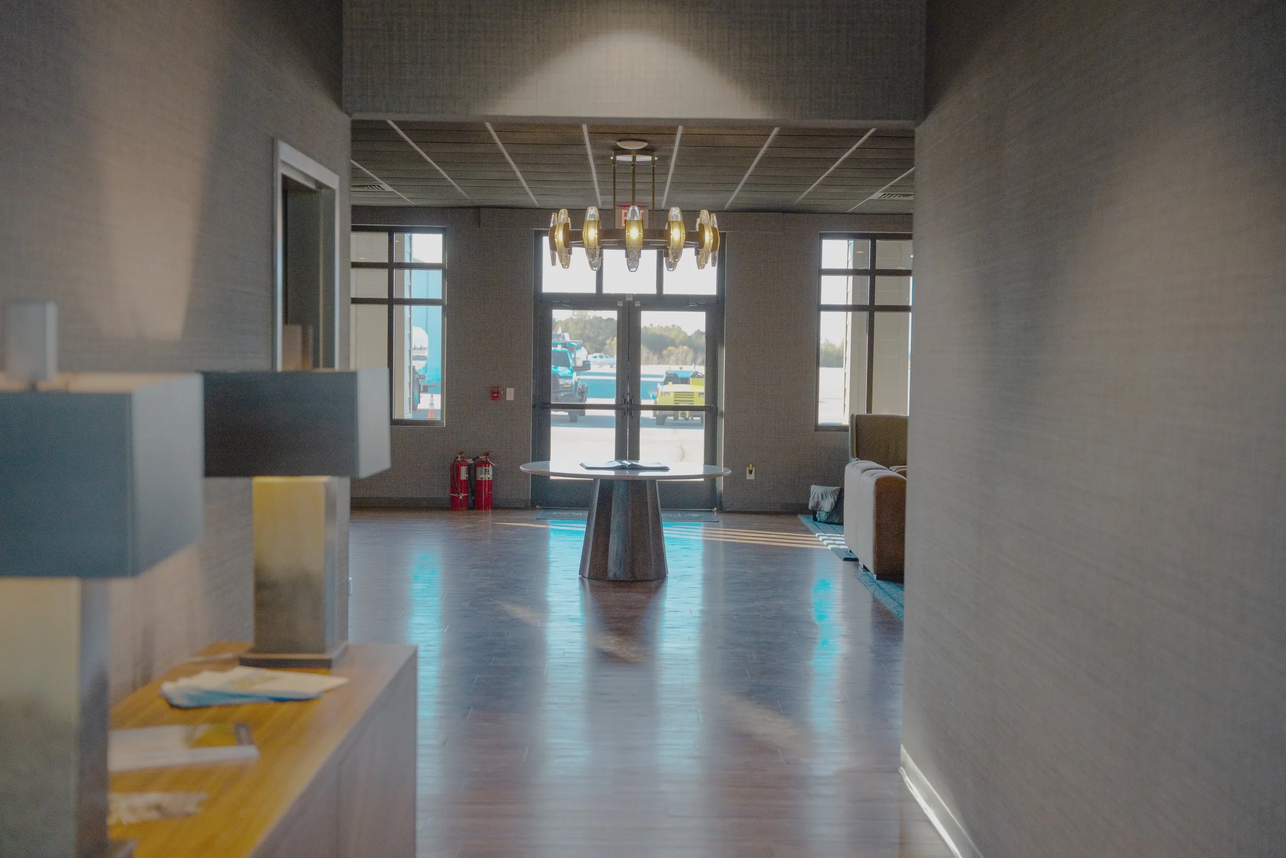 Interior of a modern room with wooden floors, a round table, a chandelier hanging from a gray ceiling, and large windows allowing natural light, with two fire extinguishers near the exit door.