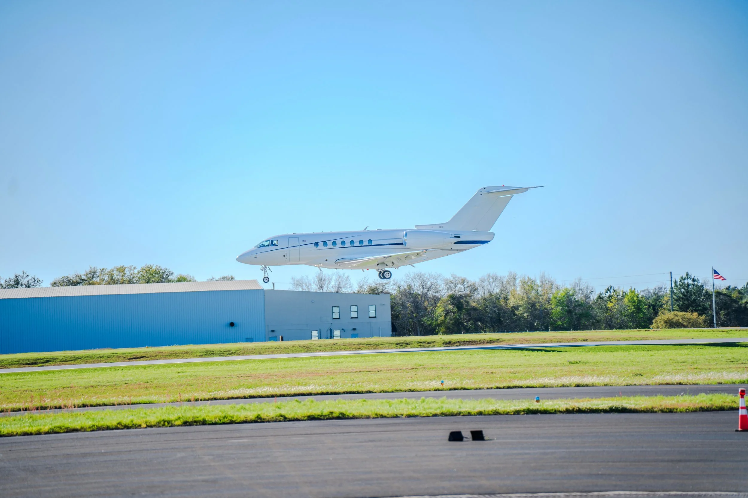 A white private jet airplane is flying low over an airport landing strip, with a hangar building and trees in the background and a blue sky.