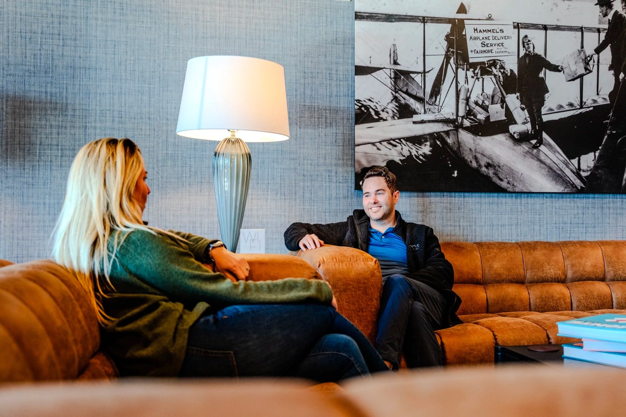 Two people sitting on a brown leather couch having a conversation in a cozy room. A white table lamp is on the left, and a black-and-white photograph of an early airplane with people working on it is on the wall behind them. There are also stacked books on a table in the foreground.
