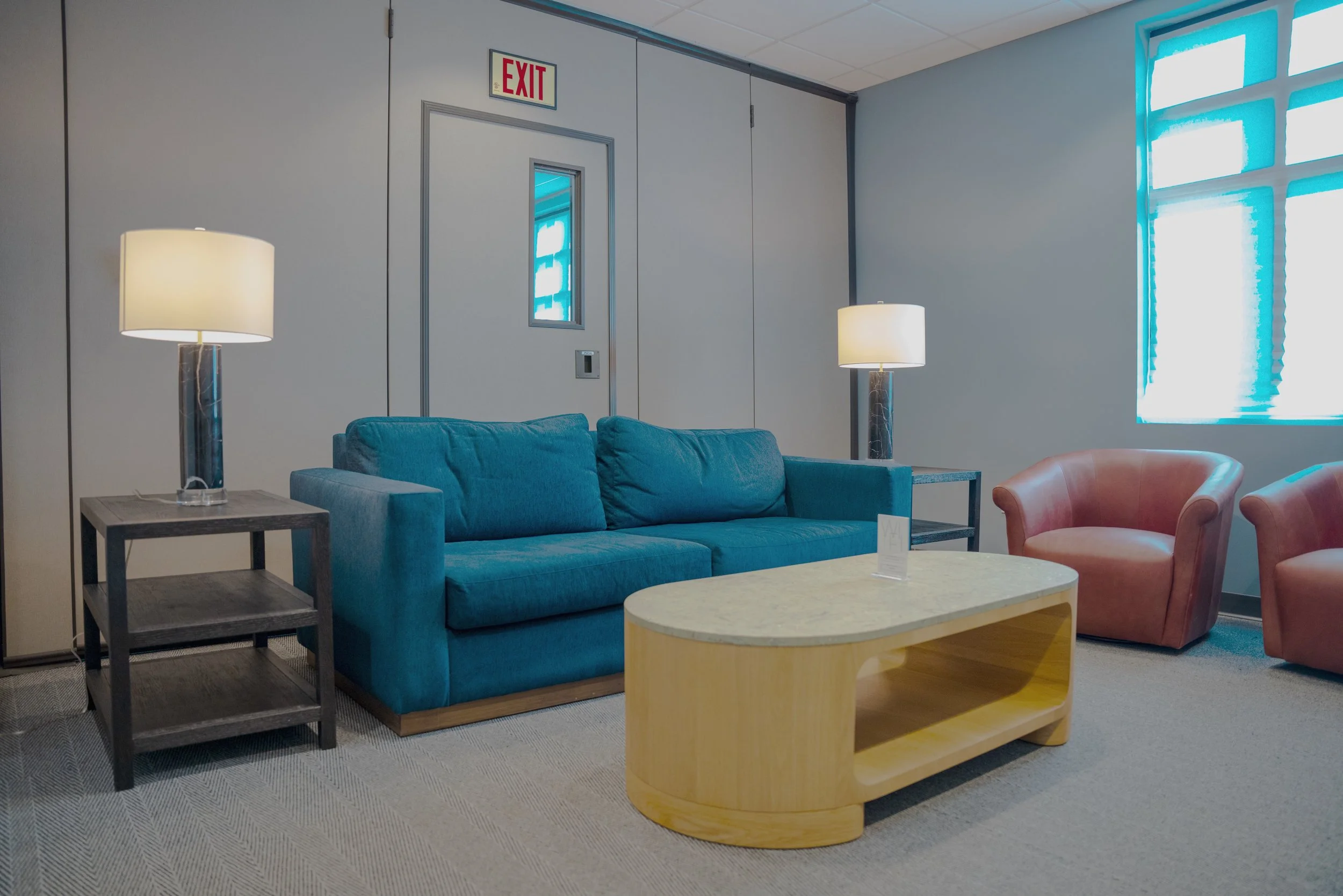 A cozy living room with a blue couch, a round wooden coffee table, two pink armchairs, and two tall black lamps on side tables. There is an exit door with a window and a framed picture of a window on the wall.