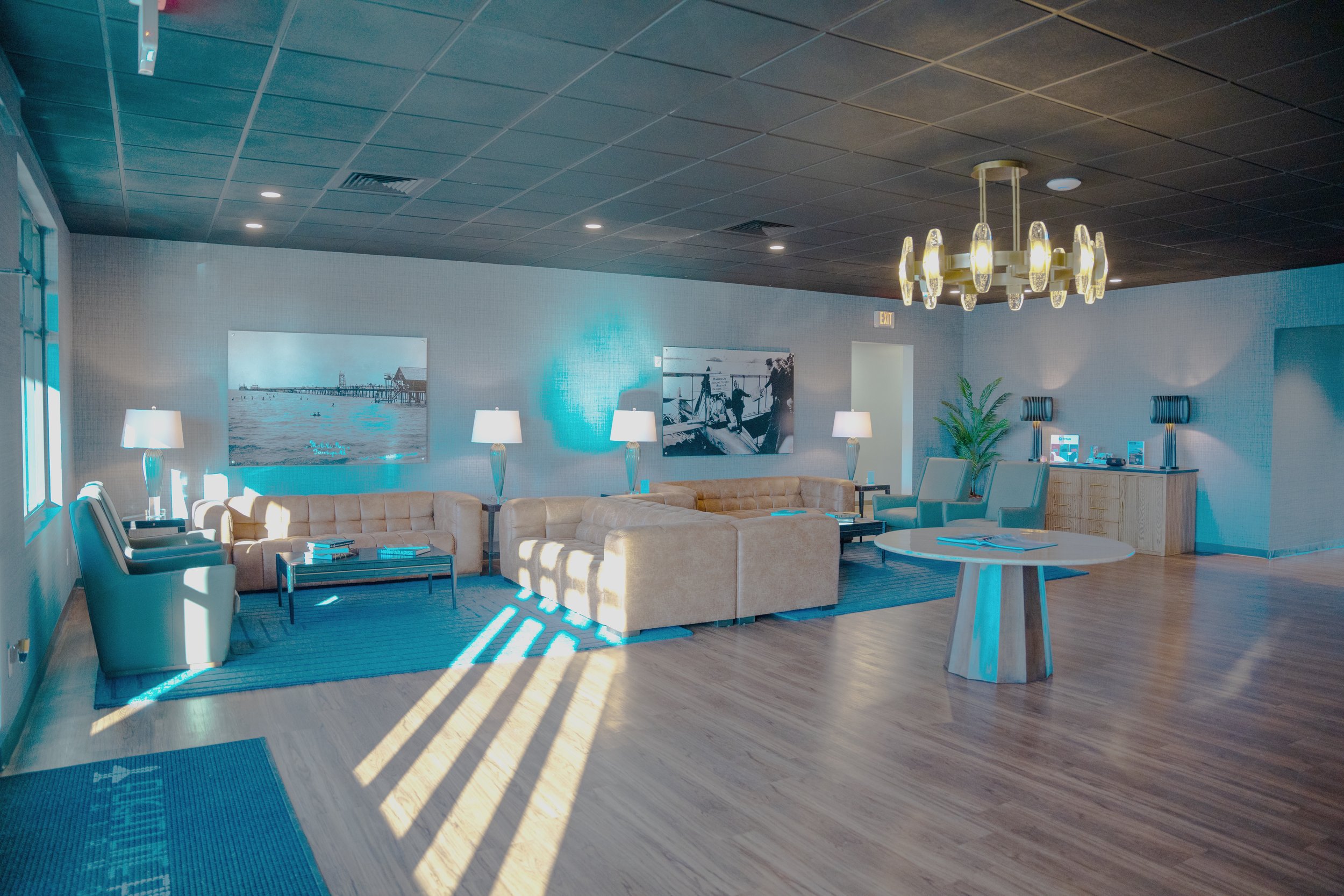 A spacious hotel lobby with light-colored sofas, chairs, and a clock on a wooden cabinet, illuminated by modern pendant lights and lamps, with artwork on the walls and a wooden floor.