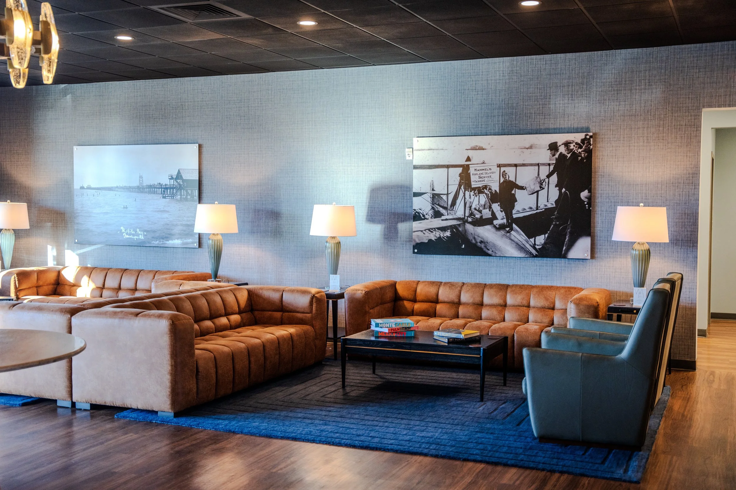 A cozy hotel lobby area with a large brown leather curved sofa, modern table lamps with white shades, a dark wood coffee table with books, and a blue armchair. The walls have framed black-and-white photographs, and the floor is wood with a rug undern