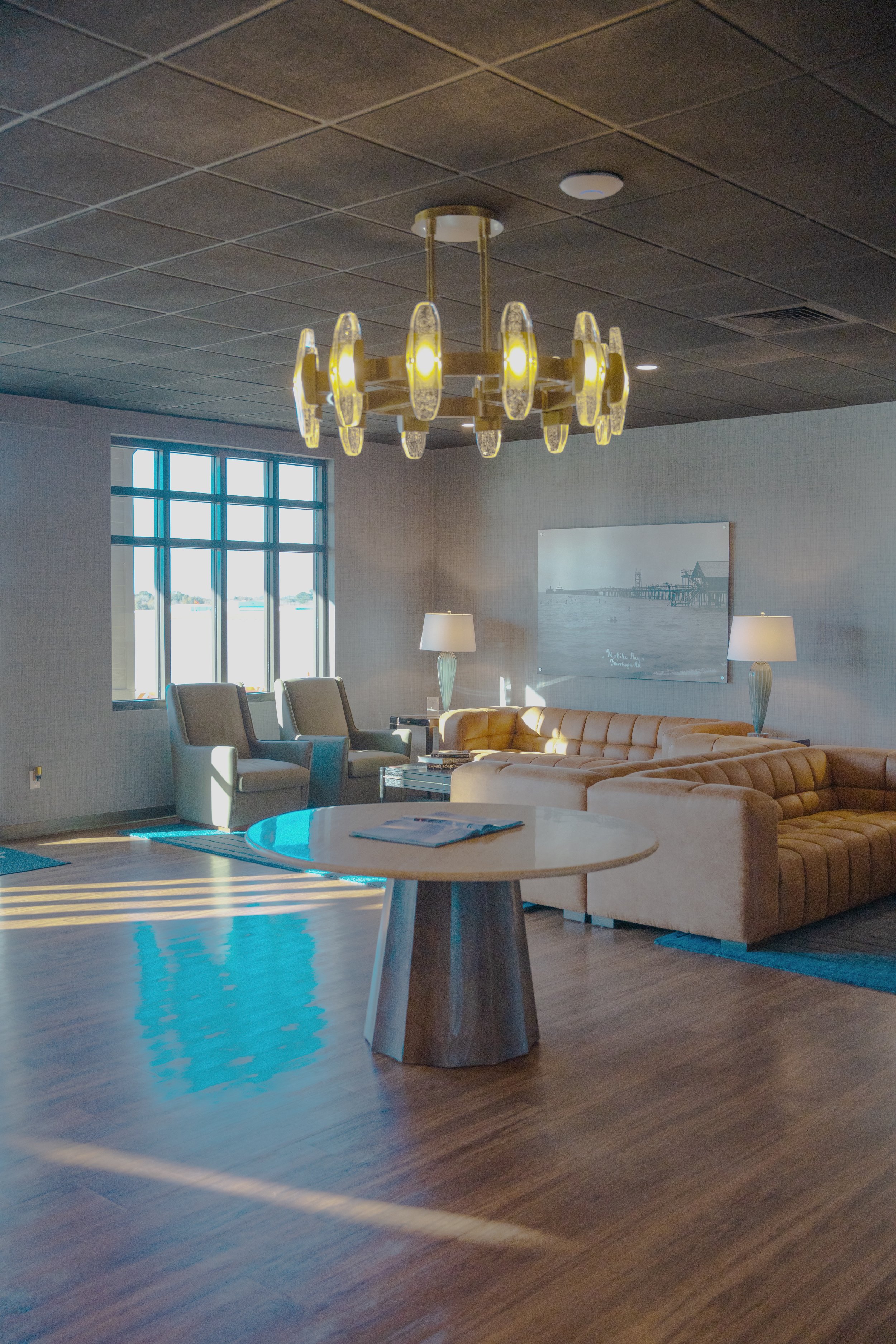A living room with a modern chandelier hanging from a black ceiling, a large window letting in natural light, beige chairs, a tan sectional sofa, a round wooden coffee table, and a side table with a tall lamp. There's artwork on the wall and a stripe
