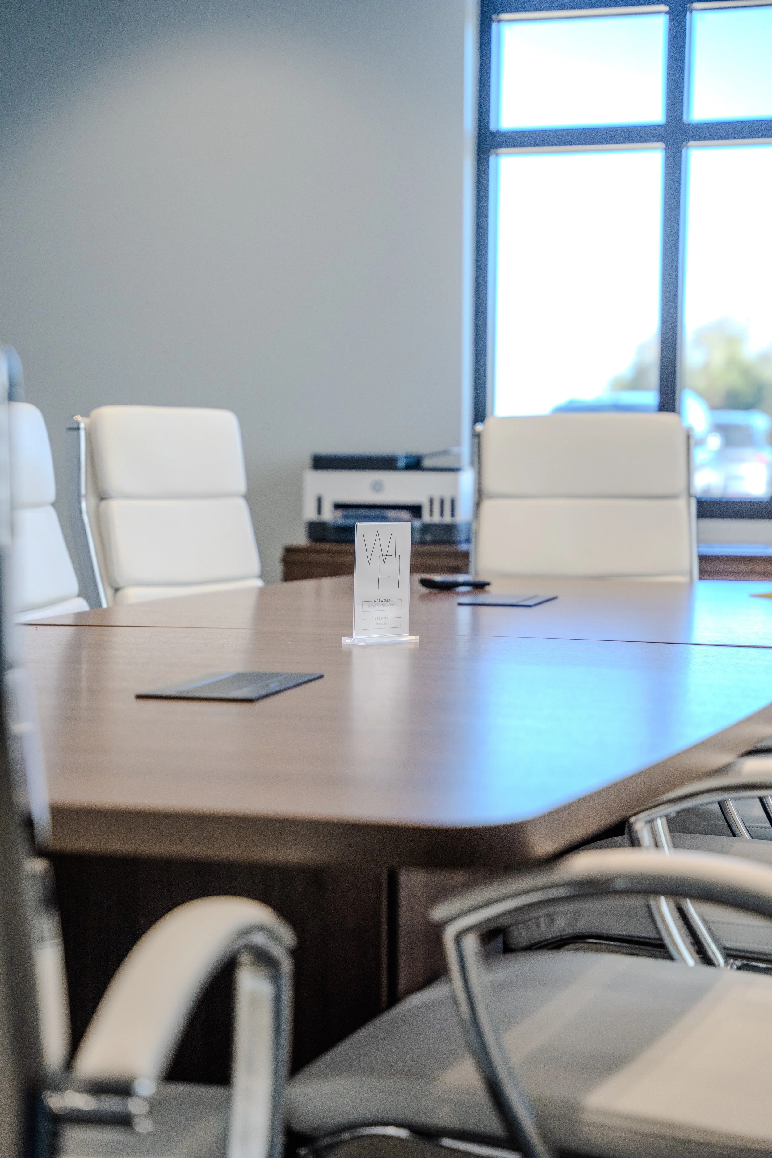 A conference room with a large wooden table surrounded by white cushioned chairs, a window letting in natural light, and a small sign on the table with text, in an office setting.