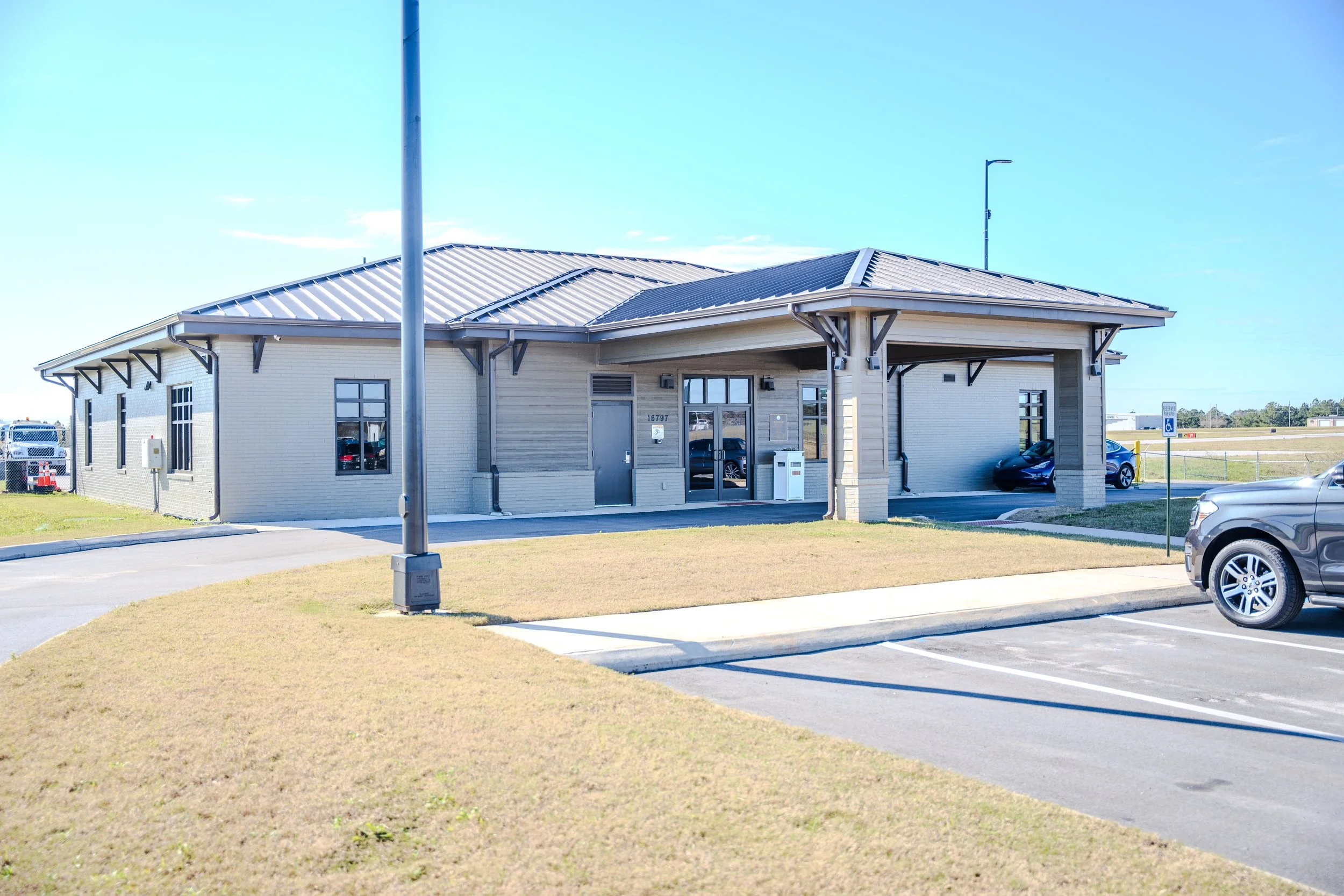 A modern building with a metal roof, glass door, and windows, surrounded by a parking lot with cars and a grassy area.