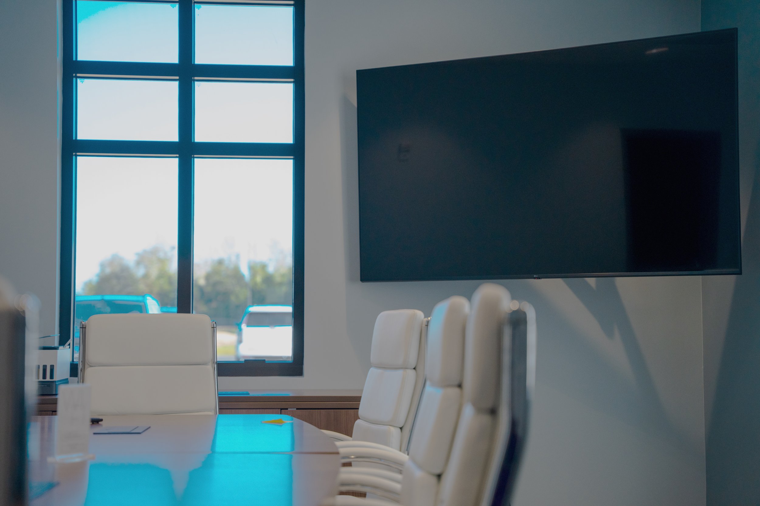 Modern conference room with a large flat-screen TV on the wall and a table surrounded by white chairs, illuminated by natural light coming through tall windows.