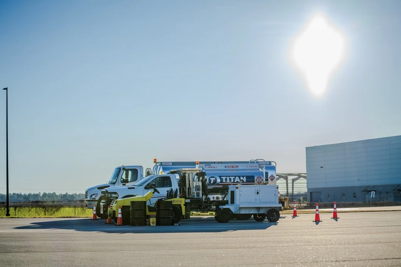 Airport fuel trucks refueling an airplane, surrounded by cones and safety barriers, with a clear sky and the sun shining brightly.
