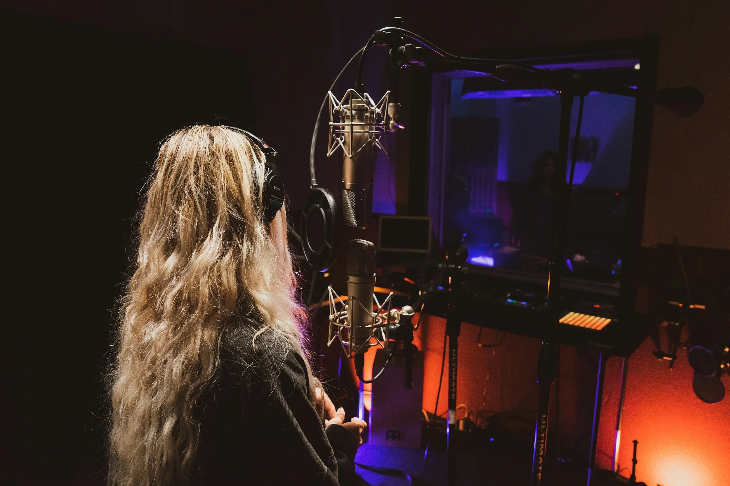 A person with long, blonde hair and headphones is in a recording studio, singing into a microphone with a pop filter, surrounded by recording equipment and a glass window showing another room with a person.