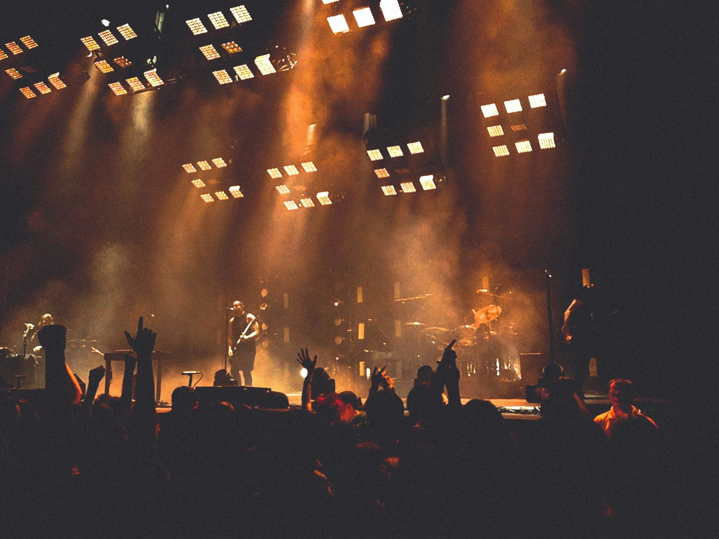 A live concert with a band performing on stage under colorful orange and yellow lights, with silhouettes of audience members raising their hands.