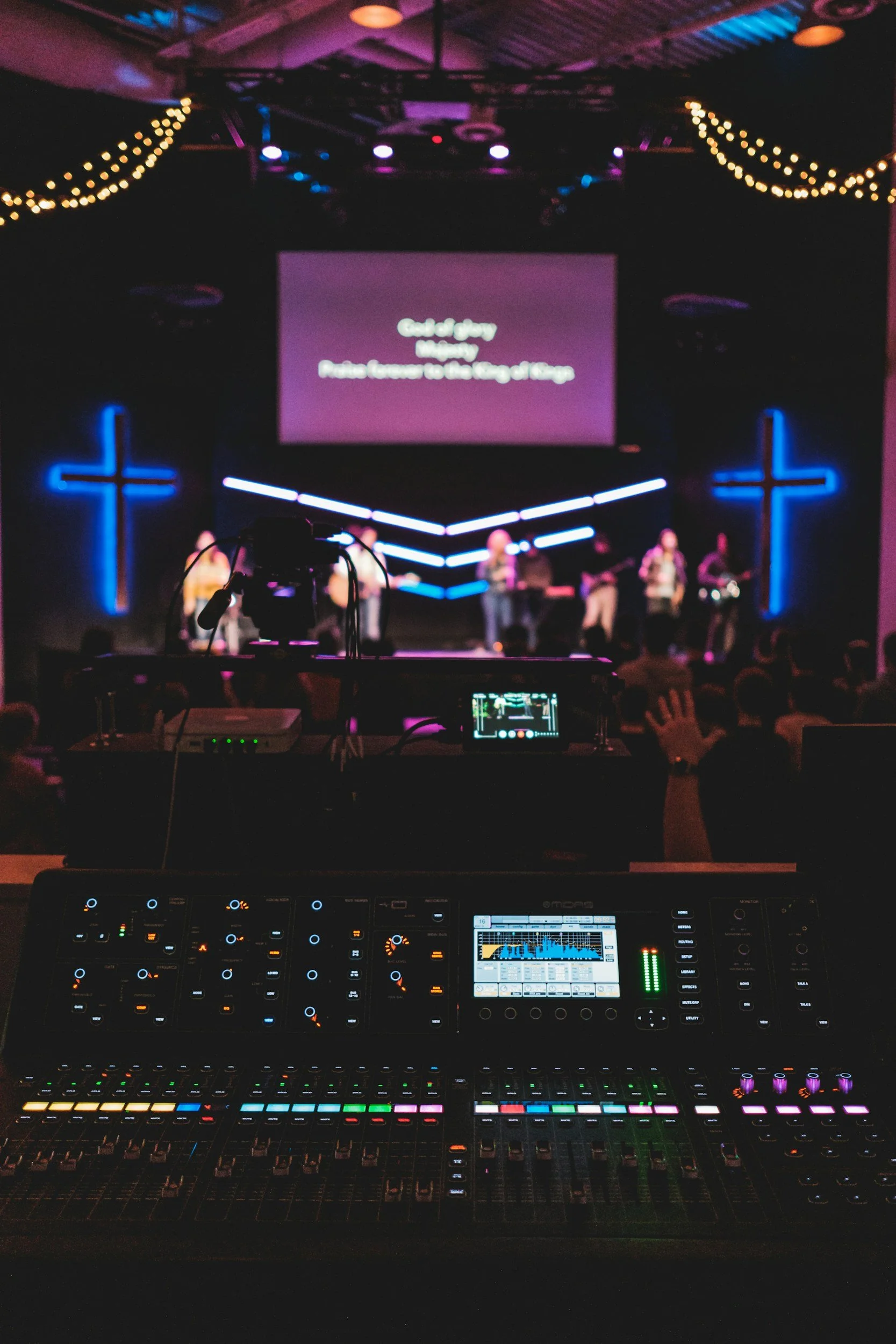A dimly lit stage with musicians performing, illuminated by blue neon cross lights. A large screen displays song lyrics, and a variety of lighting equipment is visible. In the foreground, a mixing console with numerous colorful controls and a digital screen is present, indicating a live music or worship event.