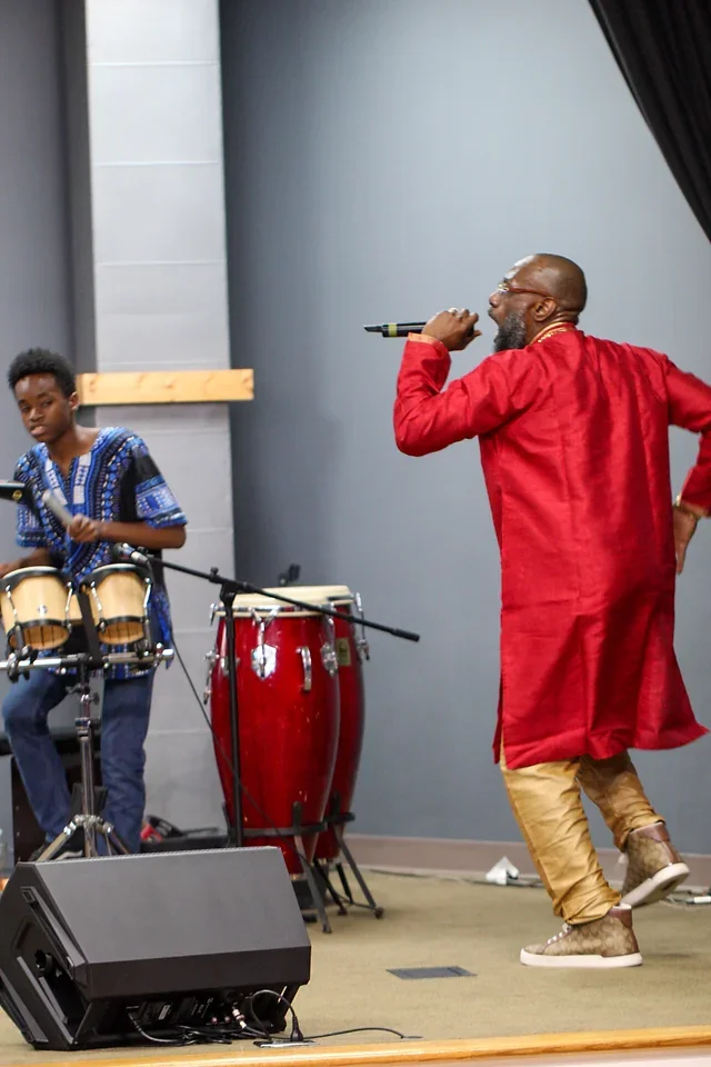 A man in a red coat and beige pants singing into a microphone with a young boy playing percussion instruments on stage, with drums and congas nearby.