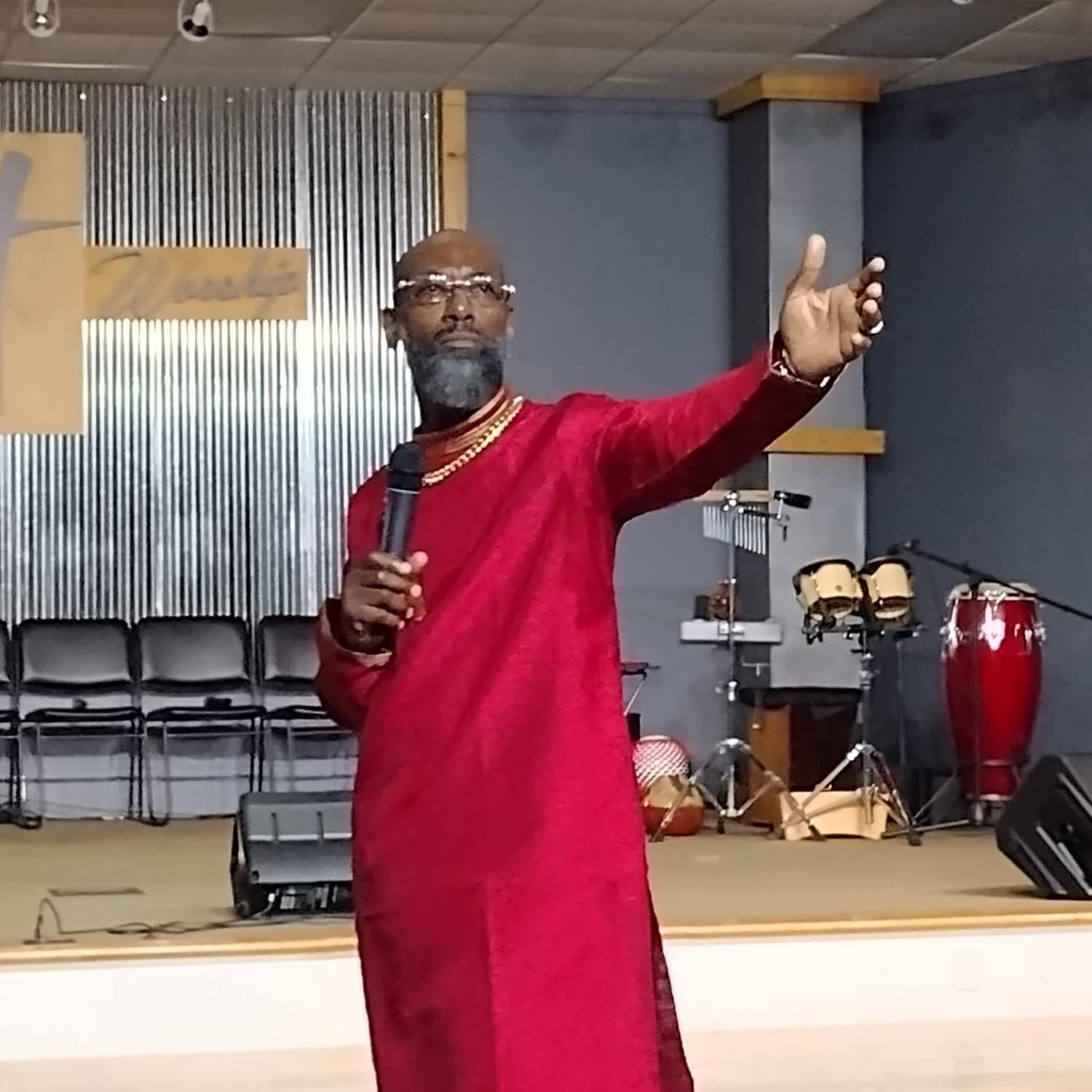 A man in a red traditional outfit holding a microphone, gesturing with his right hand, standing in a room with musical instruments in the background.