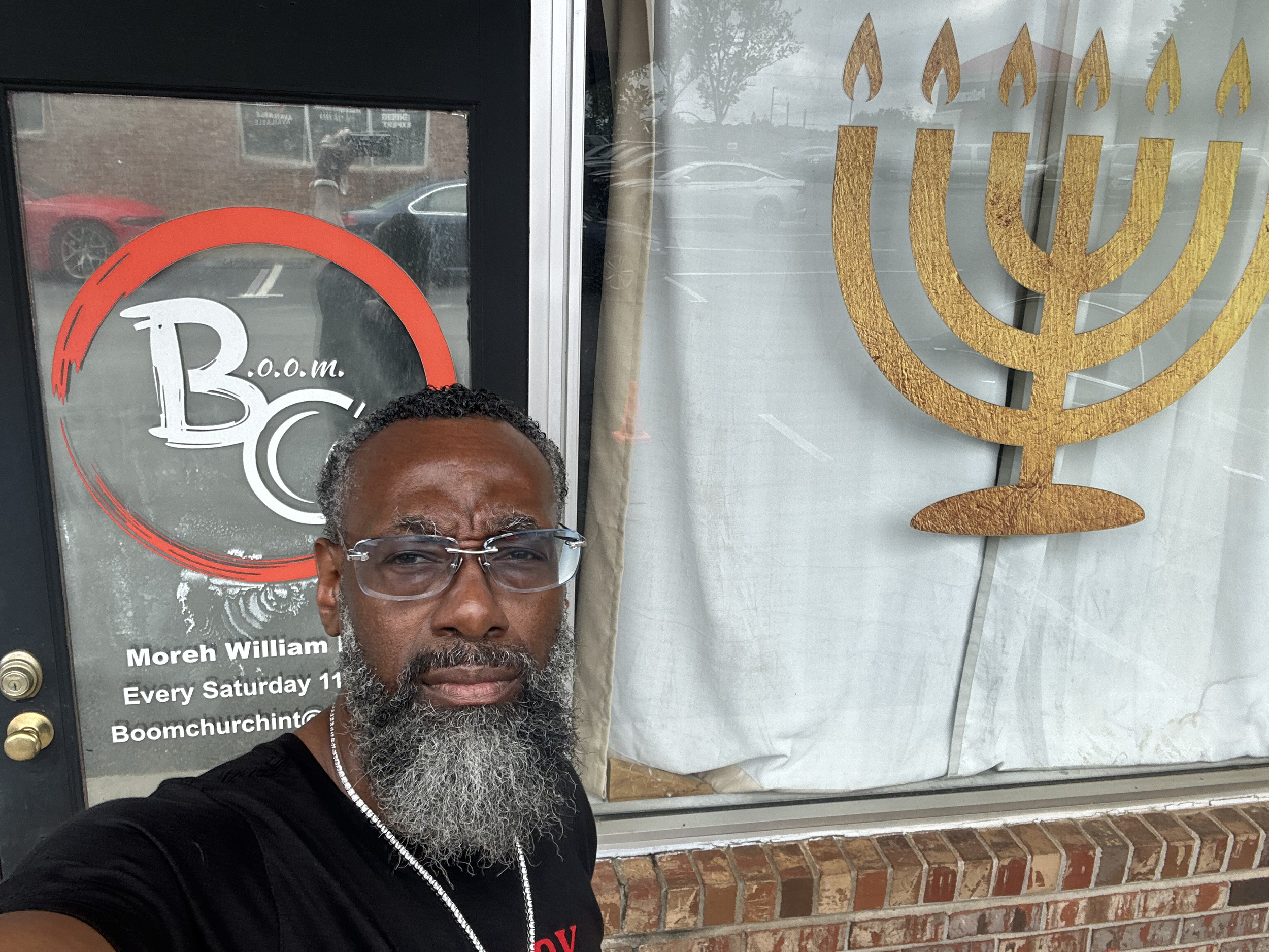 Close-up of a man with glasses and a gray beard taking a selfie outside a storefront with gold menorah and oil lamp symbols on the window. Reflection of cars and trees are visible in the window.