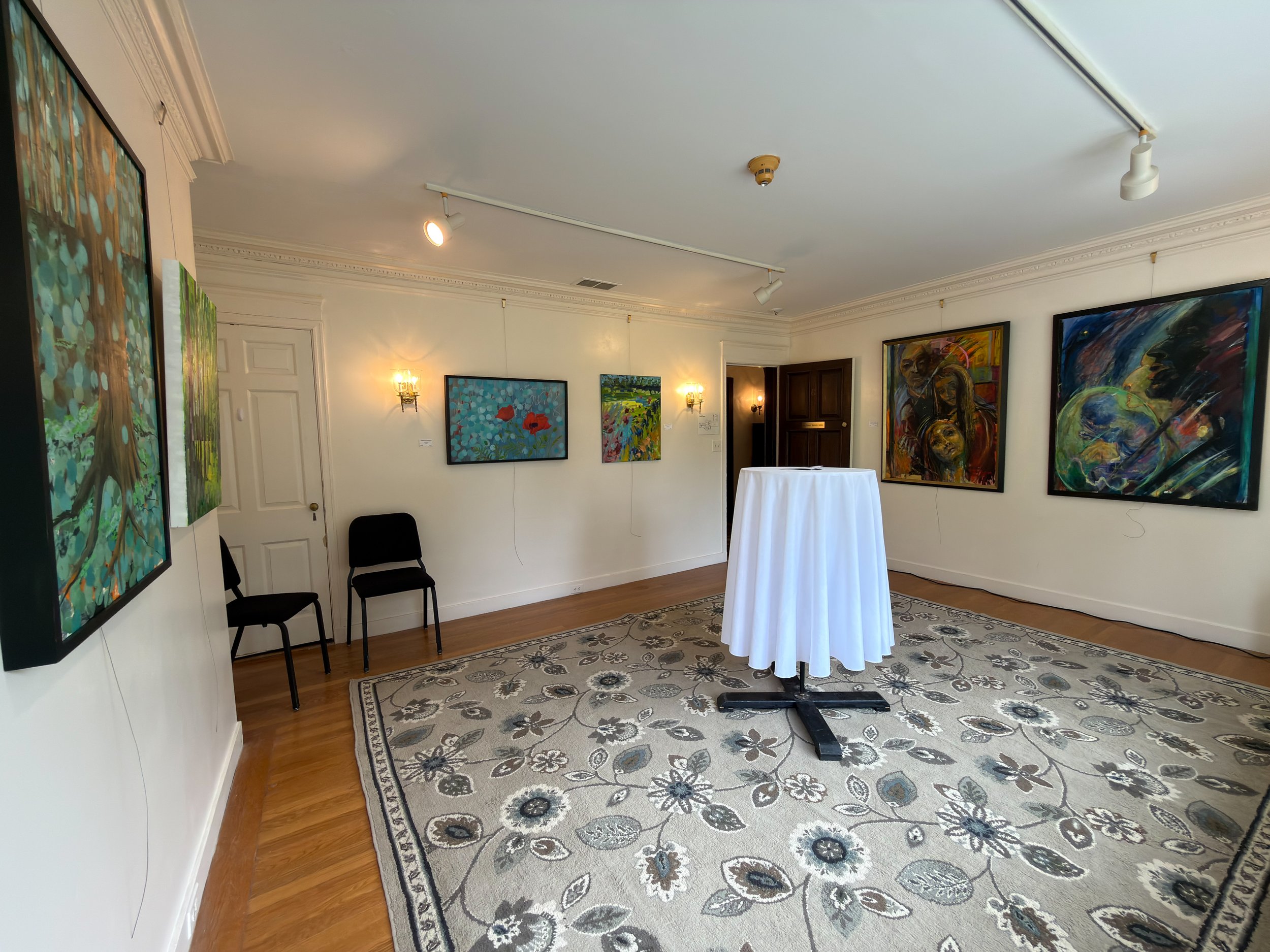 Gallery of vibrant, colorful paintings on white walls inside an art gallery with a floral-patterned rug, chairs, and a round high-top table covered with a white cloth.