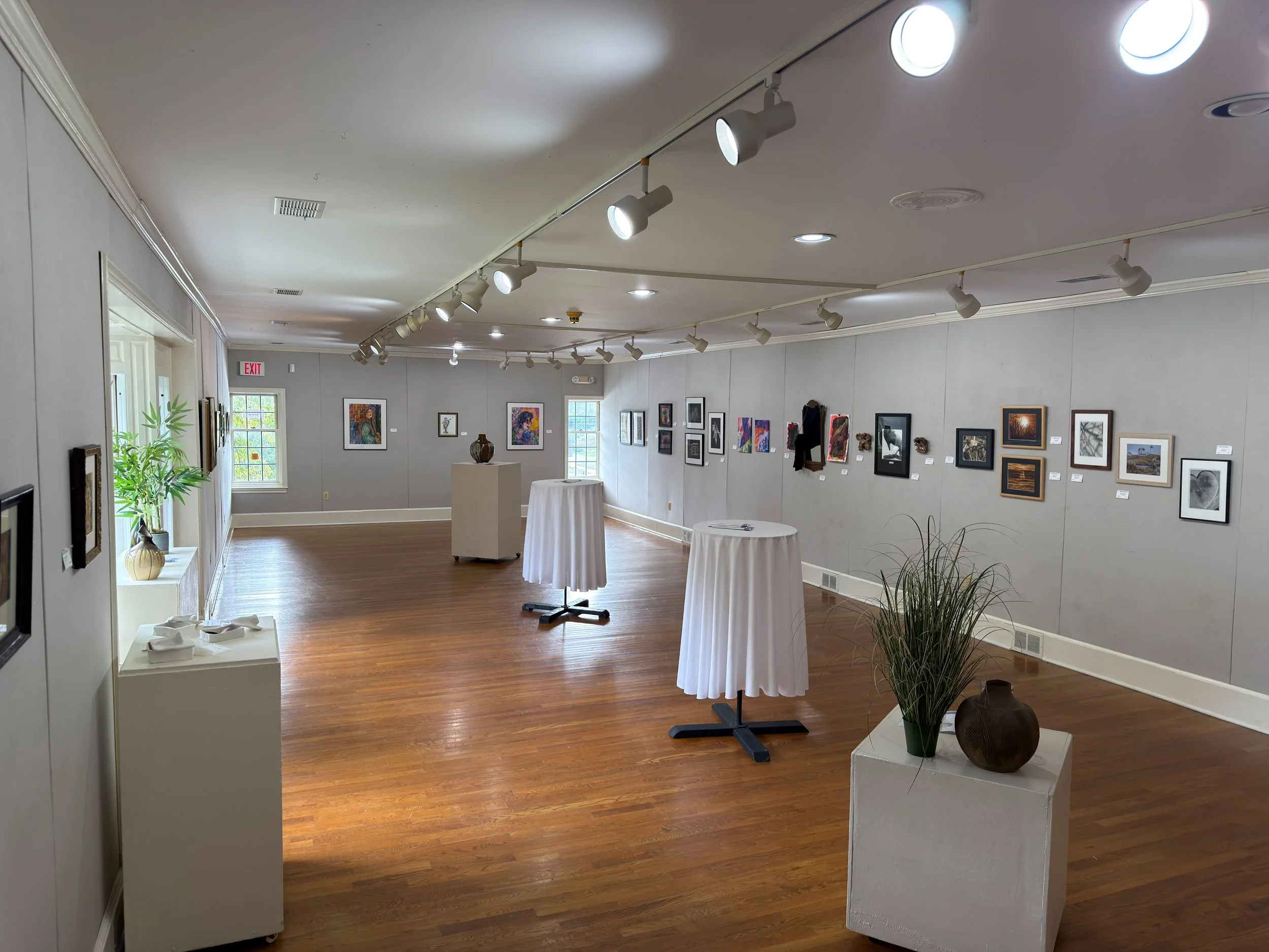 Interior of an art gallery with framed artwork on light-colored walls, spotlights on a track ceiling, and small round tables covered with white cloths, with decorative vases and a potted plant near the entrance.