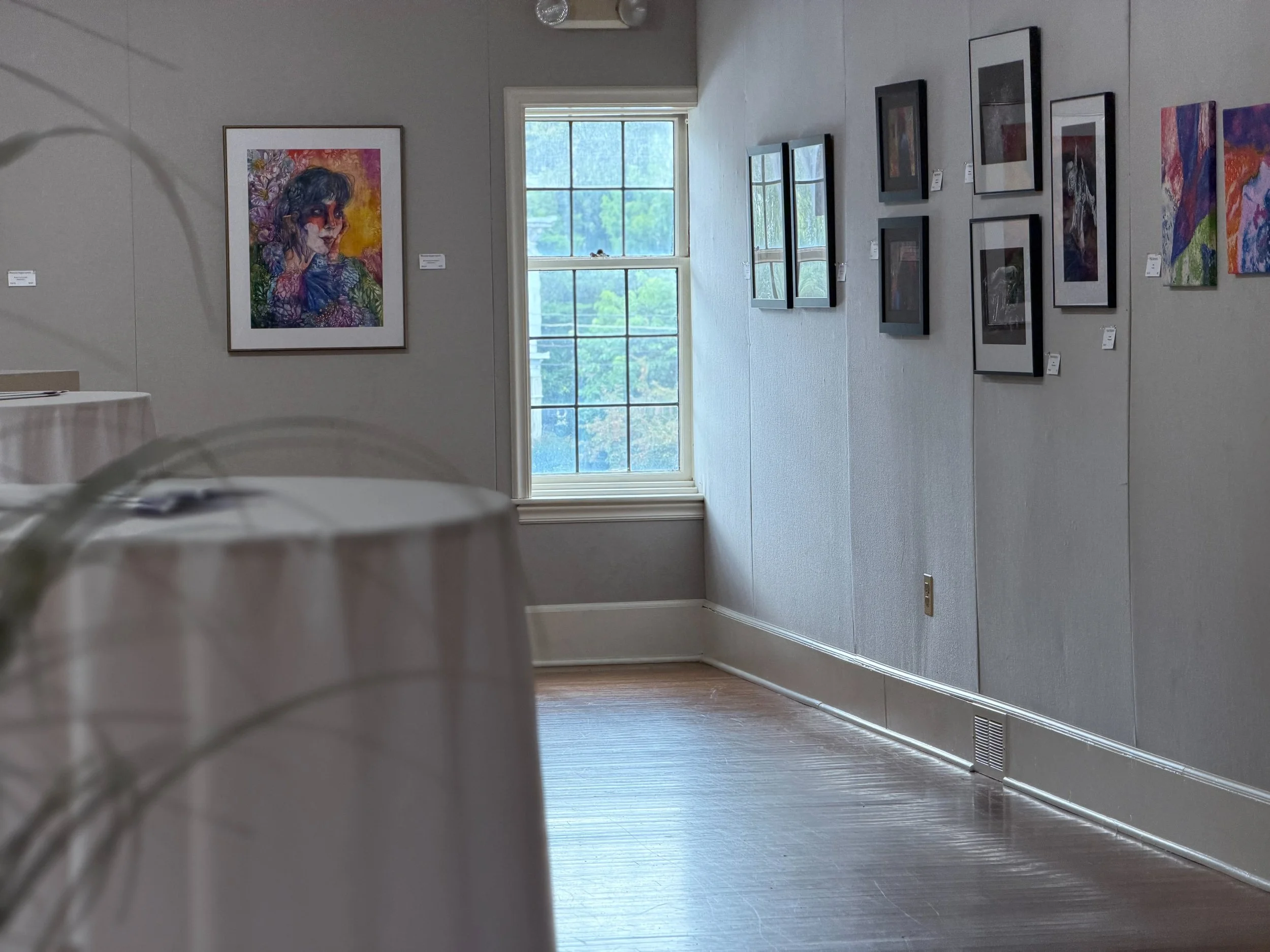 Inside an art gallery with framed artwork on the wall, a large window showing trees outside, and a wooden floor.