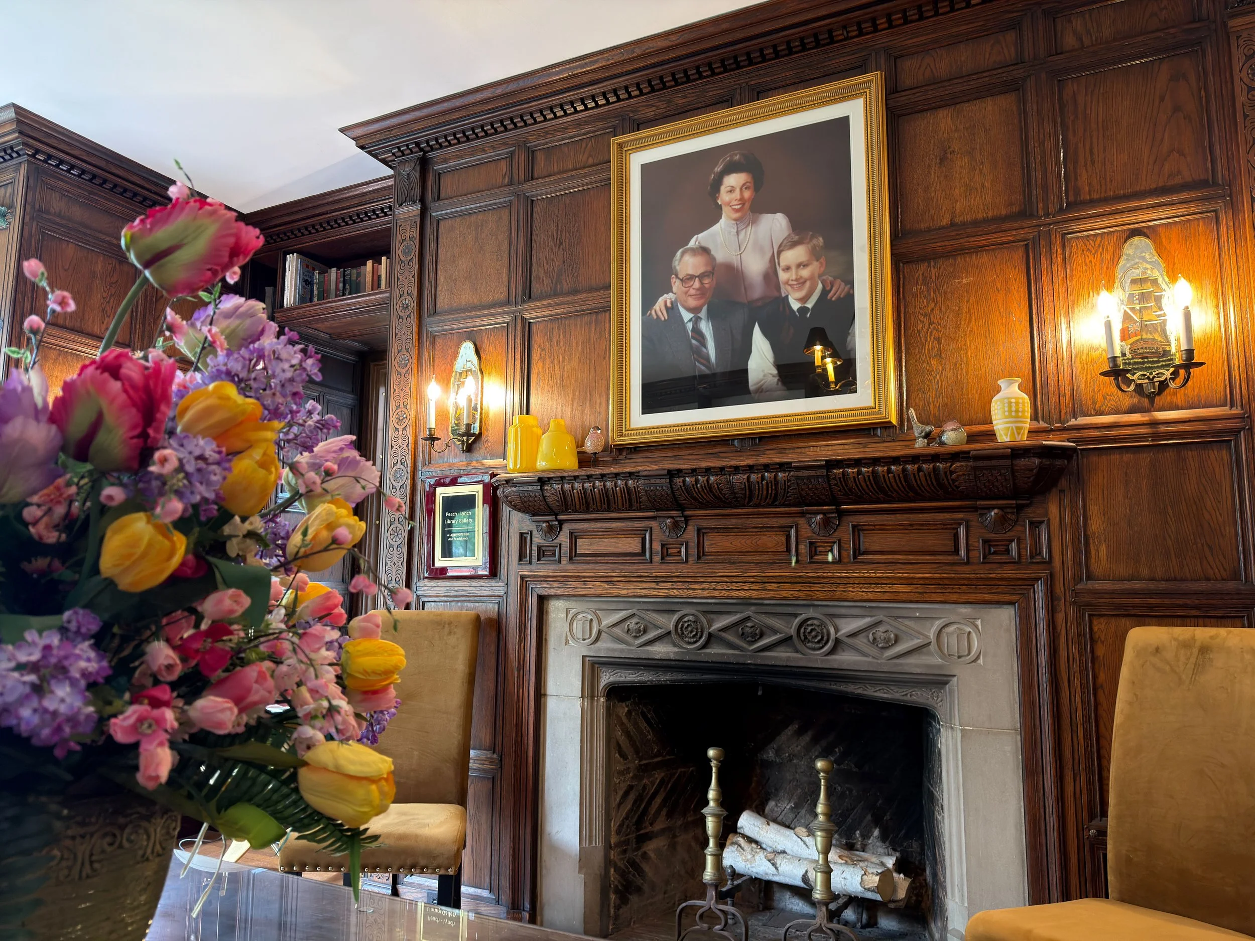 A wooden-paneled fireplace mantel with a large framed portrait of a woman and two boys hanging above it, and various decorative items including yellow vases and a small sculpture, with a bouquet of pink, yellow, and purple flowers in the foreground.