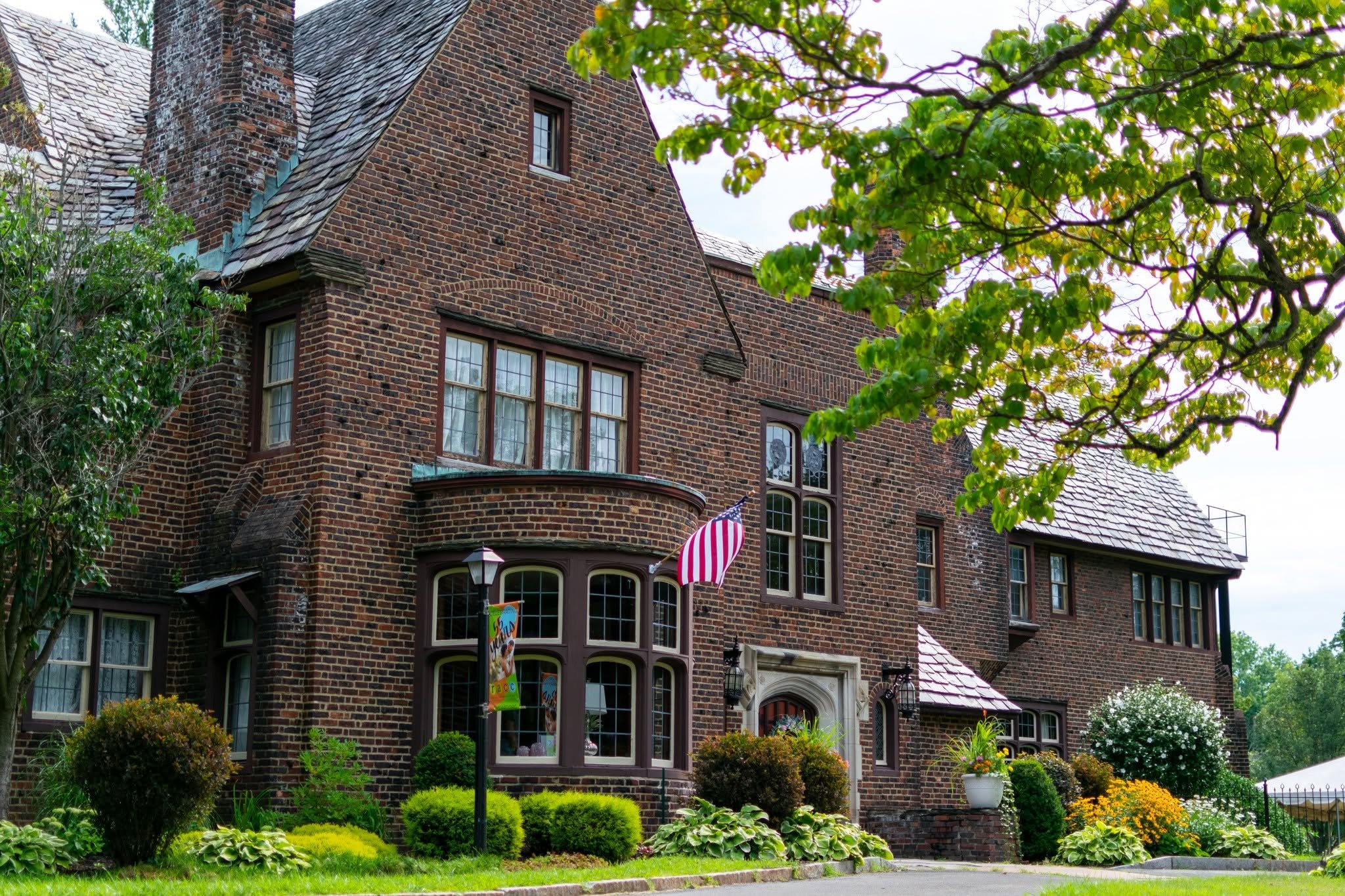 A large brick house with multiple windows and a steep roof, surrounded by green trees and a well-maintained lawn with flower bushes. There are festive decorations, including a striped red and white banner and colorful hanging ornaments, indicating a celebration or special occasion.