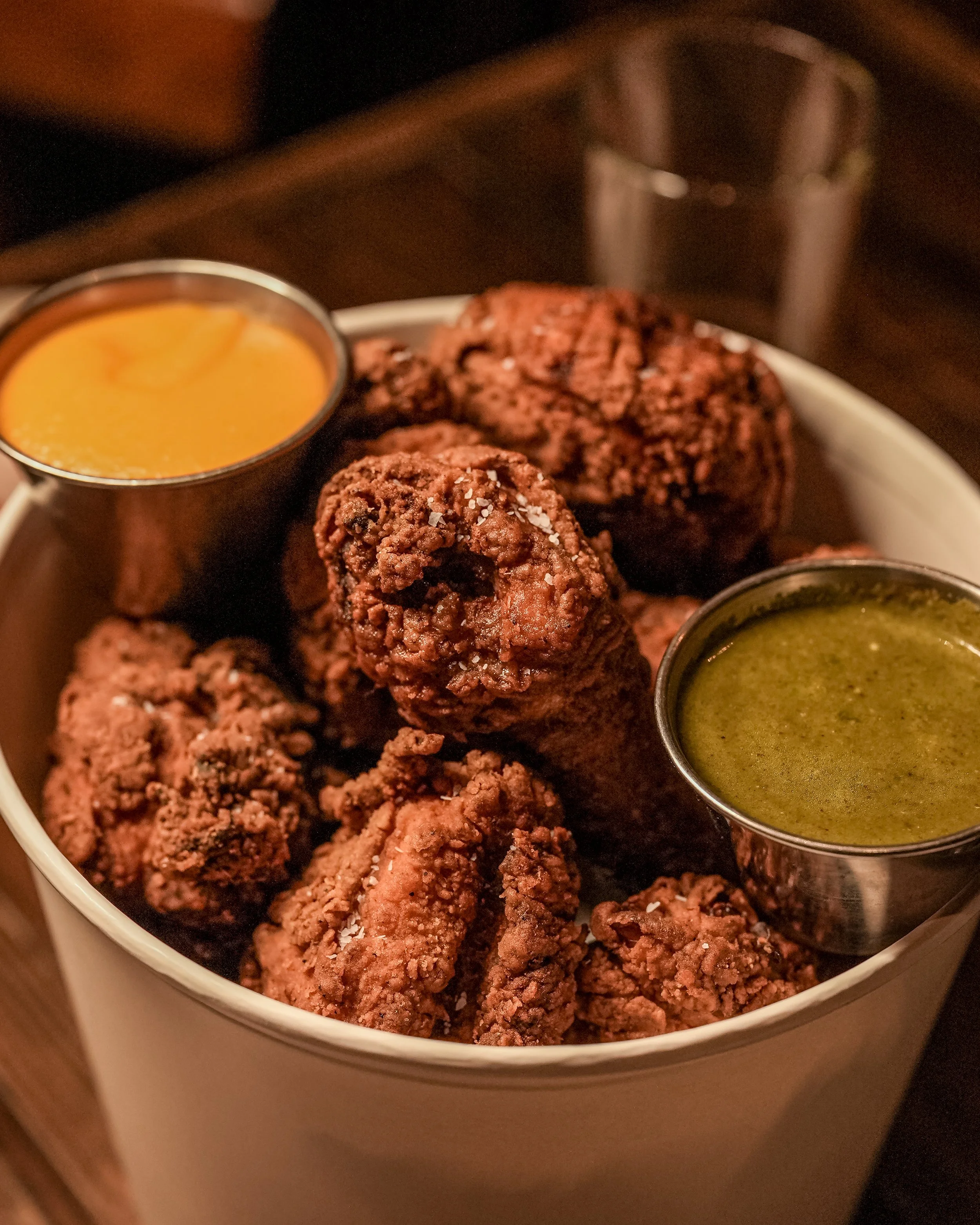 Close-up of a bowl filled with fried chicken pieces garnished with salt, accompanied by two small containers of dipping sauces, one yellow mustard and one green herb sauce.