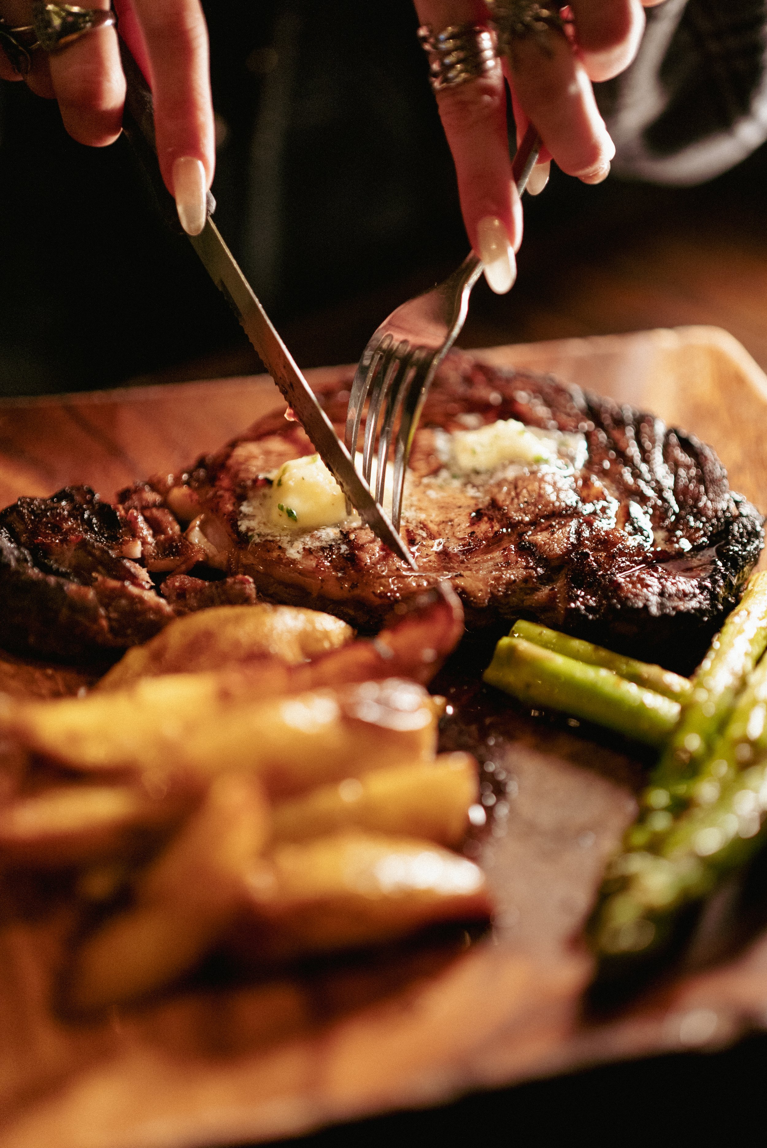 A person is carving a cooked steak on a wooden cutting board with a fork and knife. The steak has dollops of herb butter on top and is accompanied by roasted potatoes and green beans.