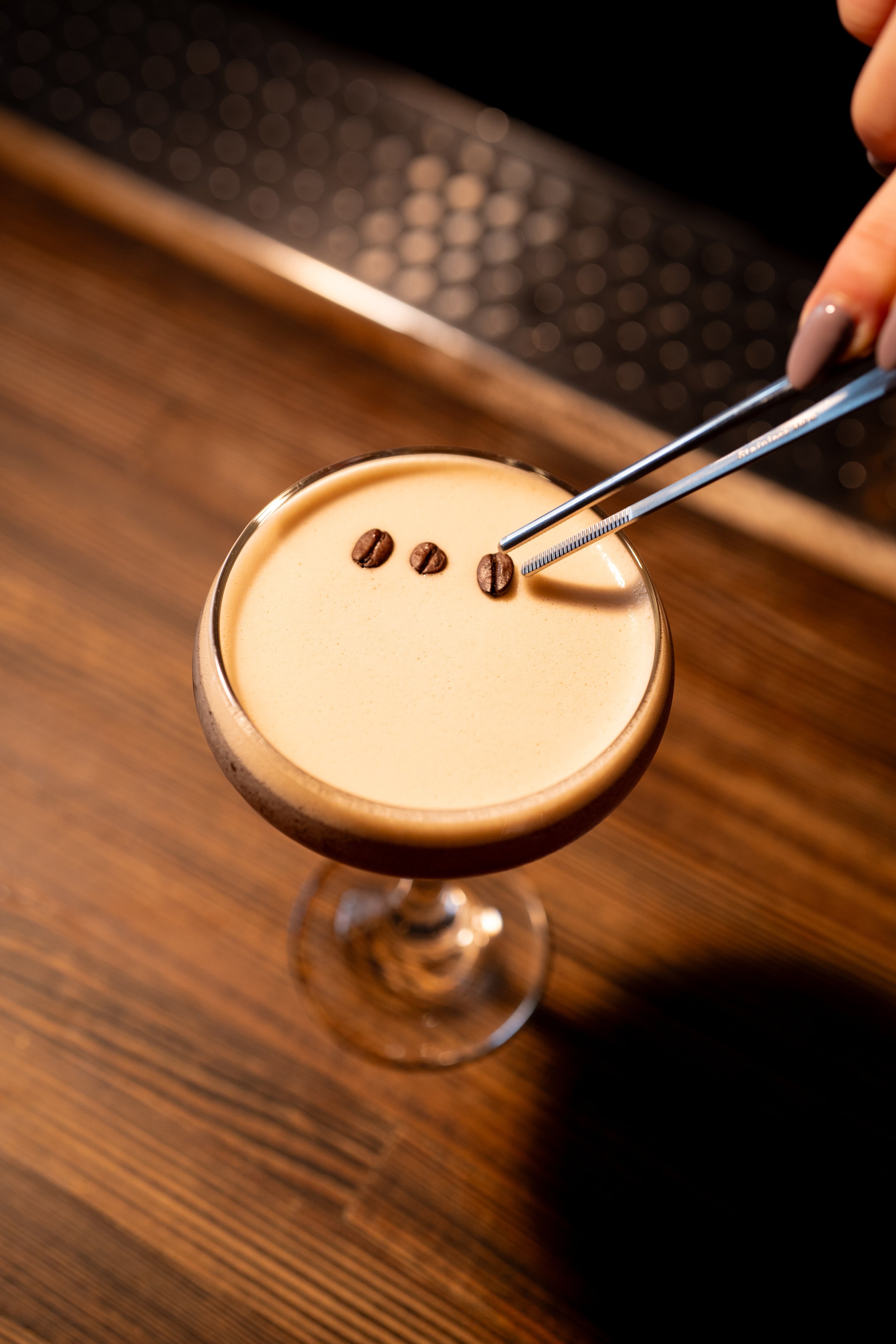 A close-up of a cocktail glass filled with a creamy coffee-colored drink, garnished with three coffee beans on top, with tweezers placing an additional coffee bean, on a wooden surface.