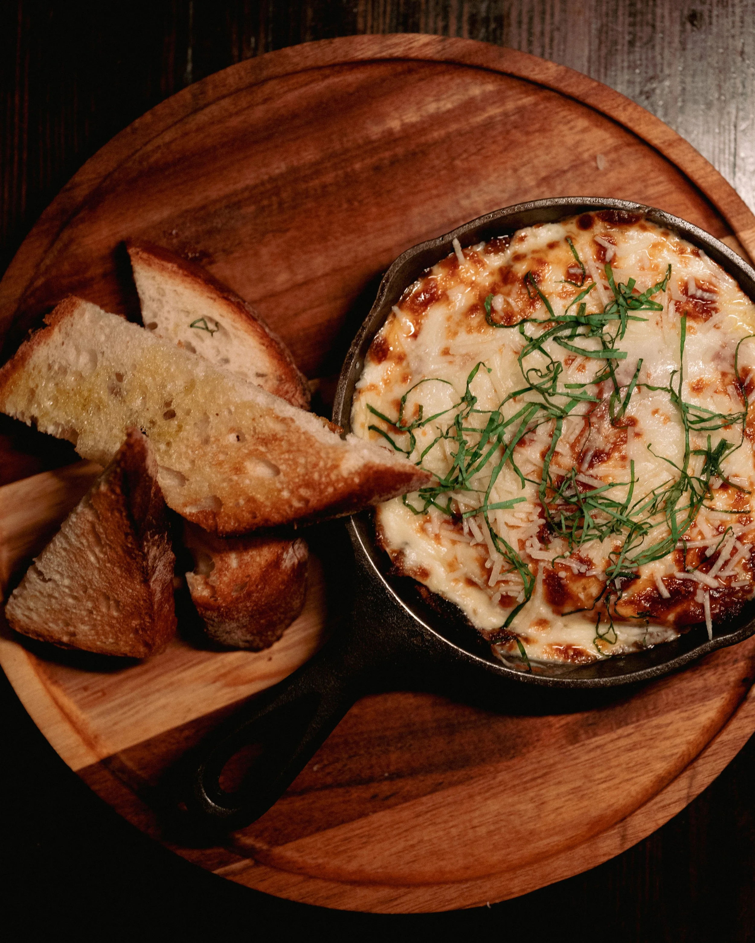 A wooden serving board with slices of toasted bread and a small cast iron dish of baked cheesy dip garnished with chopped herbs.