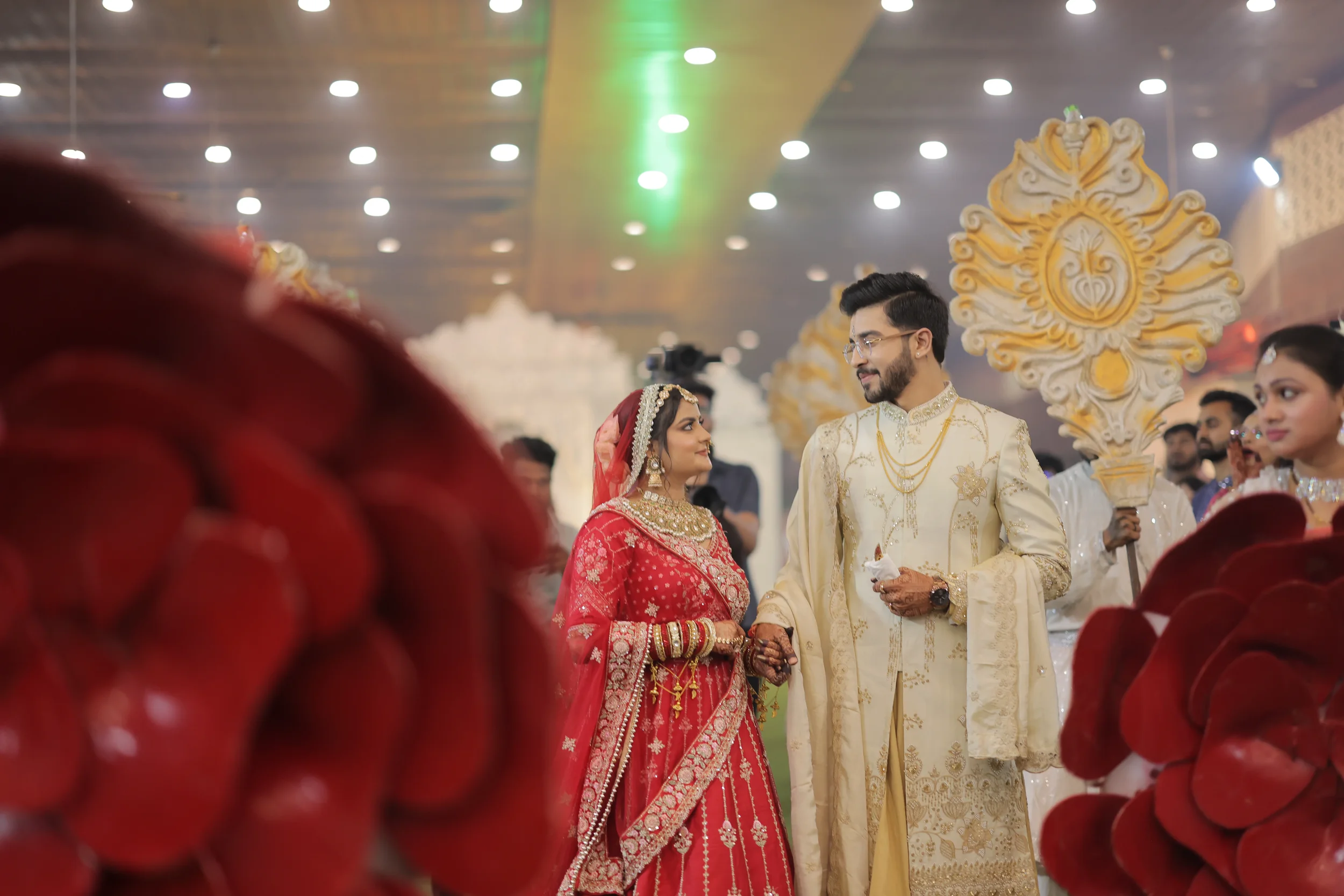 A bride and groom dressed in traditional Indian wedding attire holding hands during a wedding ceremony, with wedding guests in the background and decorative floral arrangements.