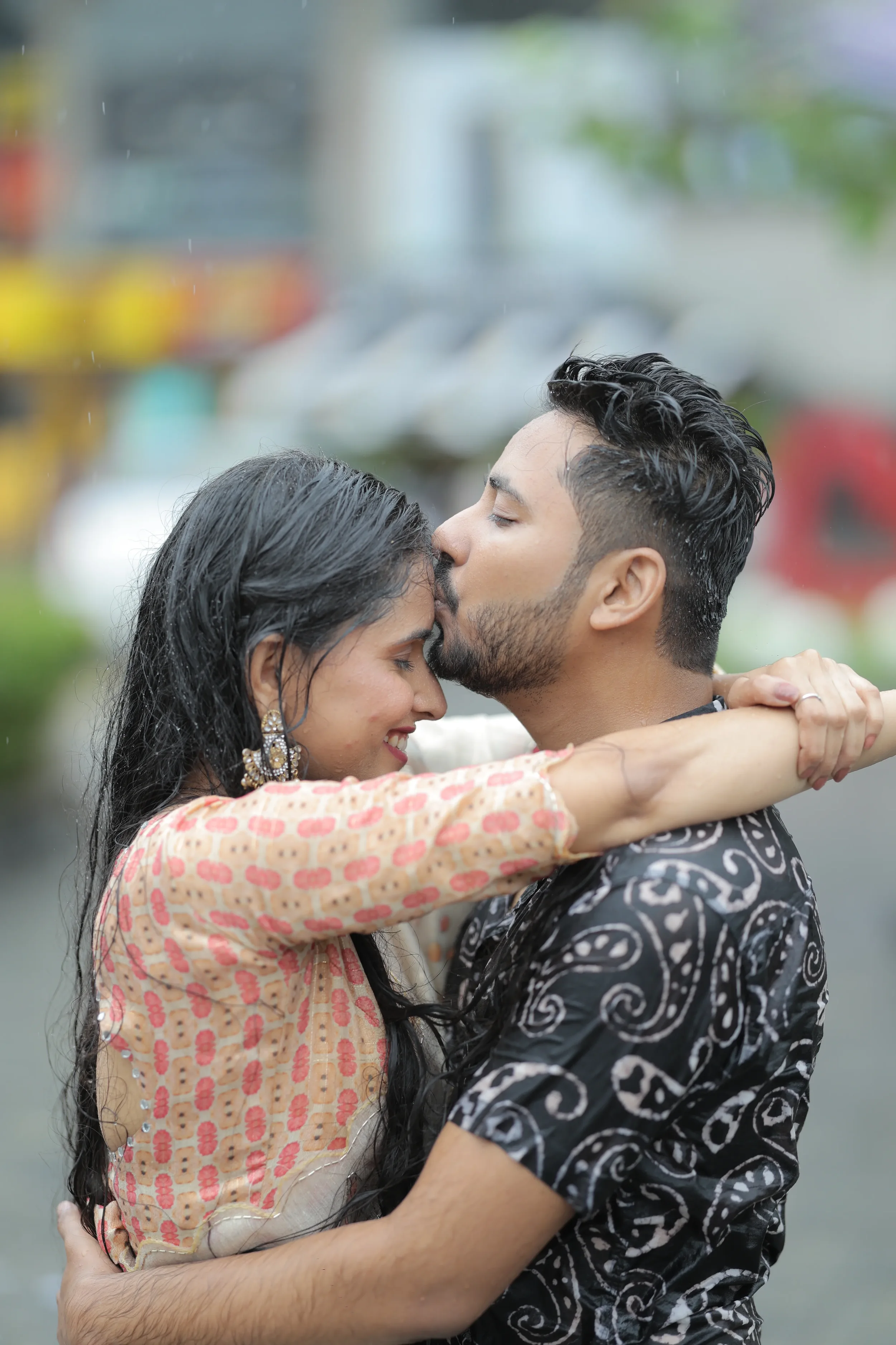 A young couple sharing a kiss outdoors in the rain. The woman has long, dark, wet hair and wears earrings and traditional Indian clothing. The man has short, wet hair and a beard, and wears a patterned shirt.