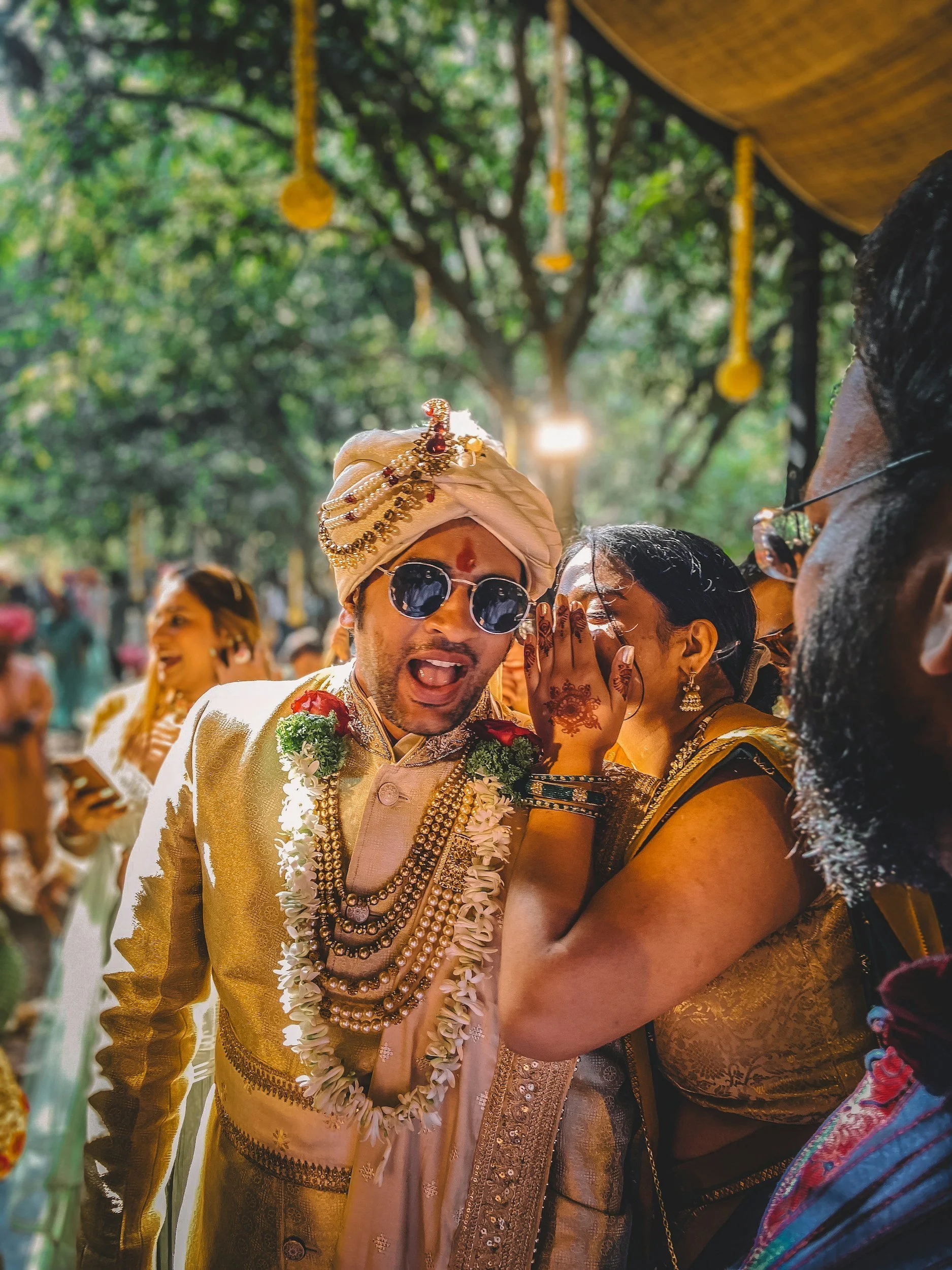 Indian groom wearing traditional attire and jewelry, with a turban and sunglasses, at a wedding celebration with guests in the background.