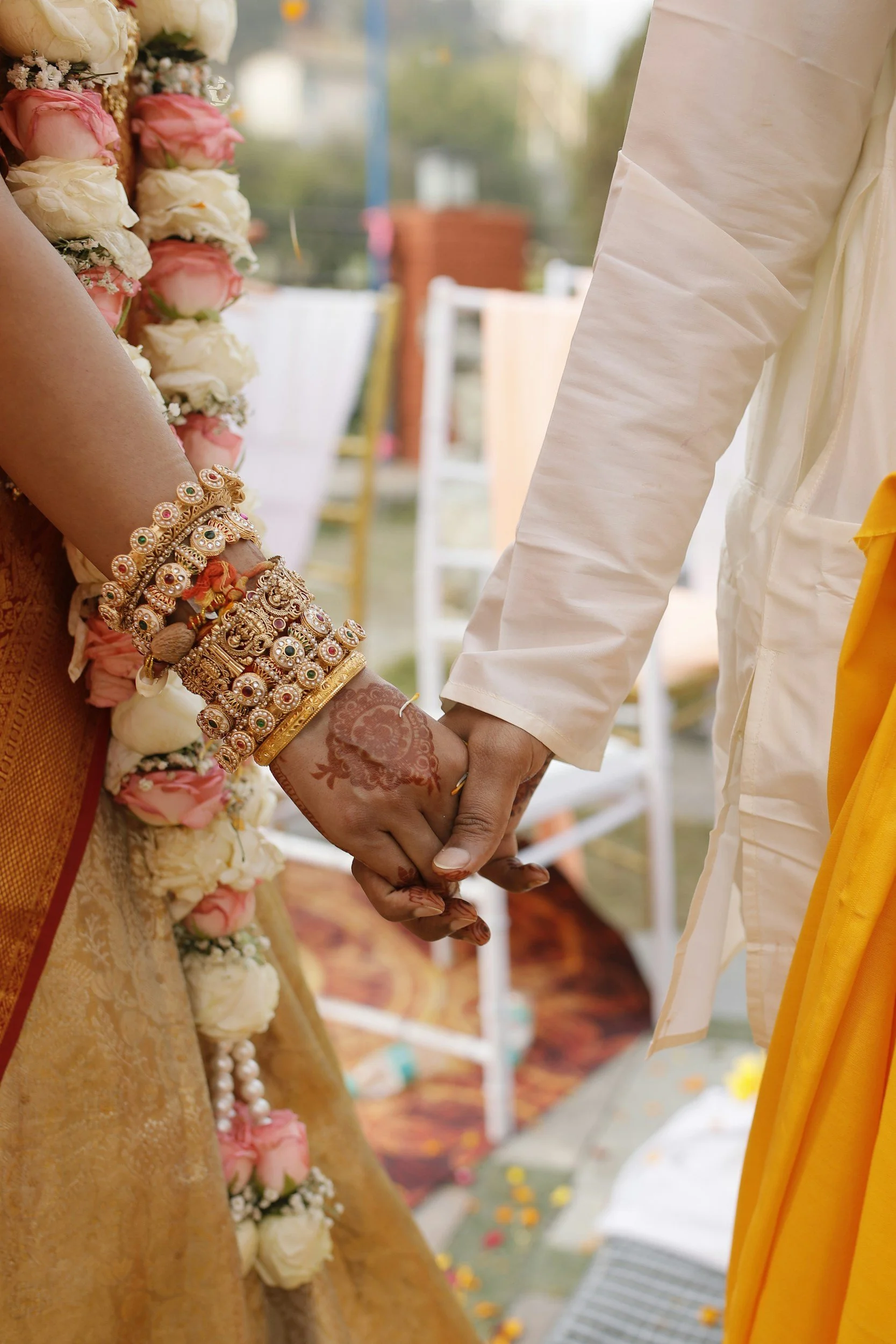 A close-up of a bride and groom holding hands during a wedding ceremony, with the bride wearing ornate jewelry and floral garlands, and the groom dressed in traditional attire.