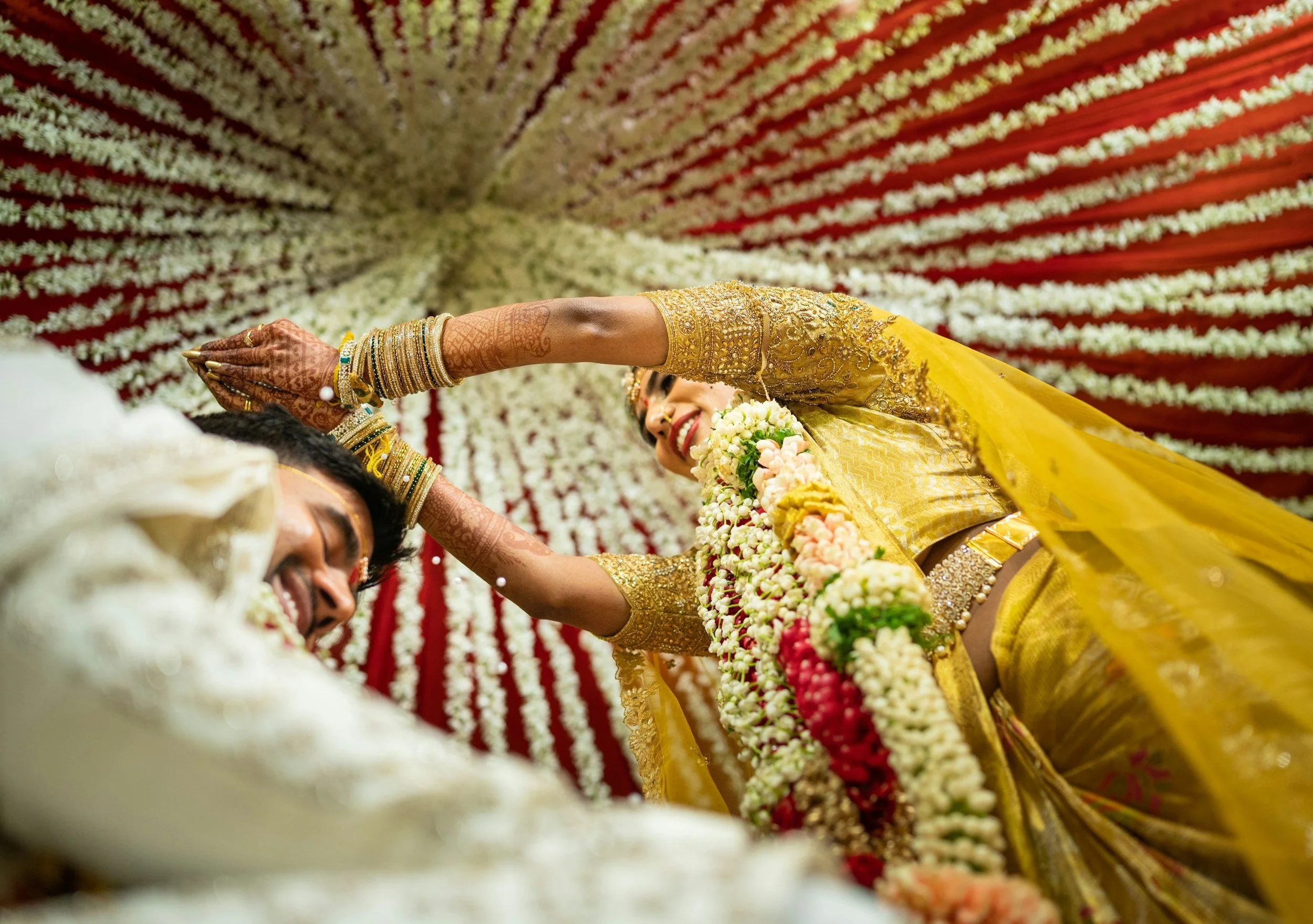 A bride dressed in a yellow sari and traditional jewelry, smiling and touching the forehead of a groom during a wedding ceremony, with decorative floral backdrop.