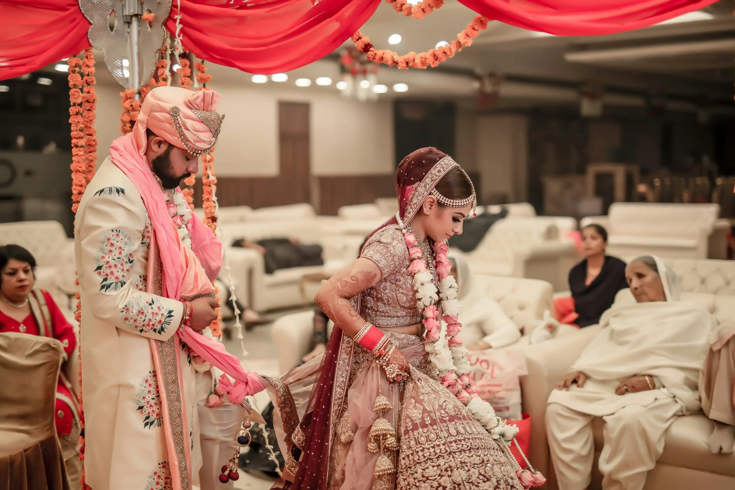 A bride and groom dressed in traditional Indian wedding attire, bowing their heads during a ceremony under pink drapery and floral decorations surrounded by family and friends.