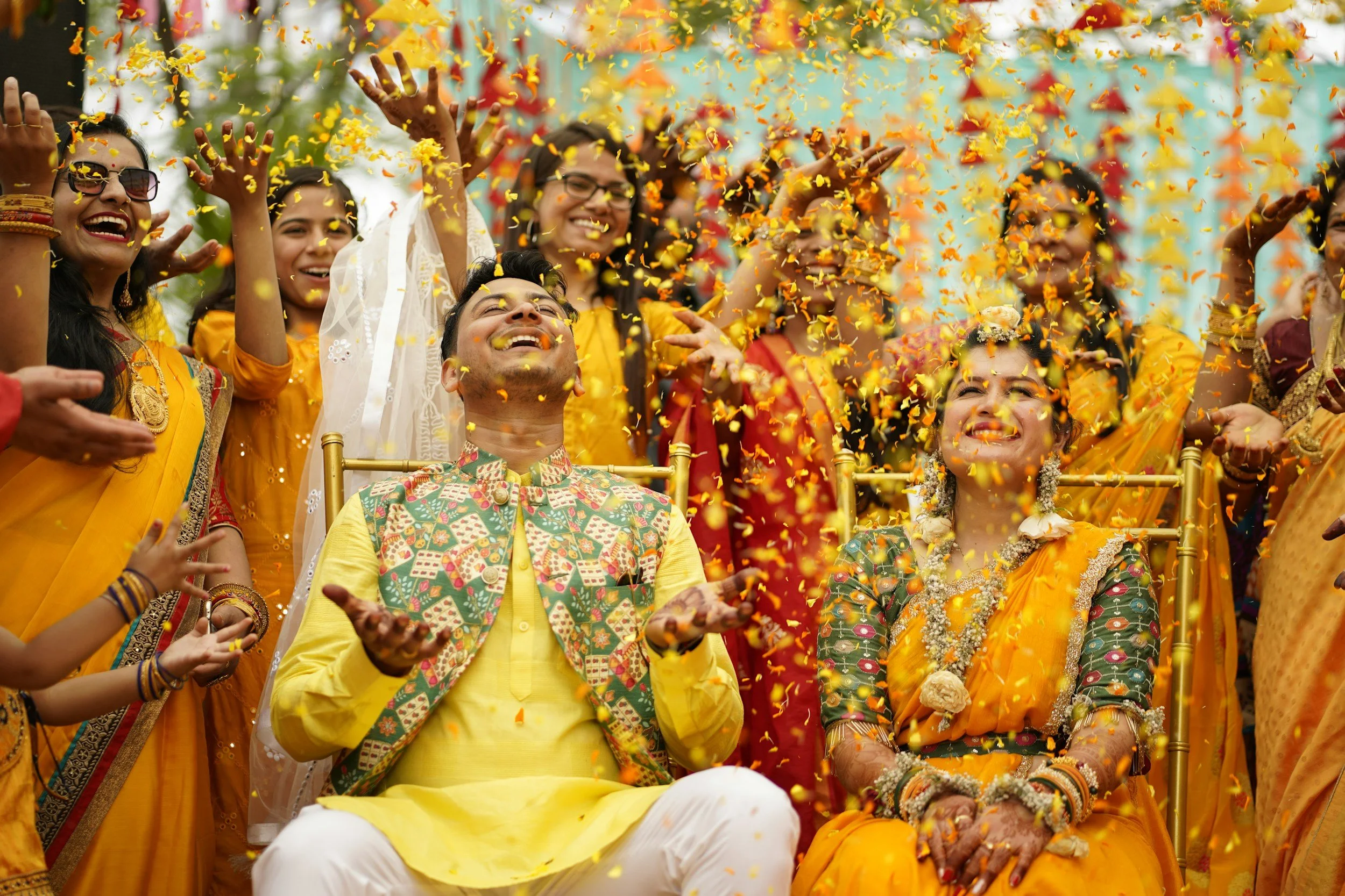 A joyful group of people celebrating with colorful flower petals in the air, dressed in vibrant traditional Indian attire.