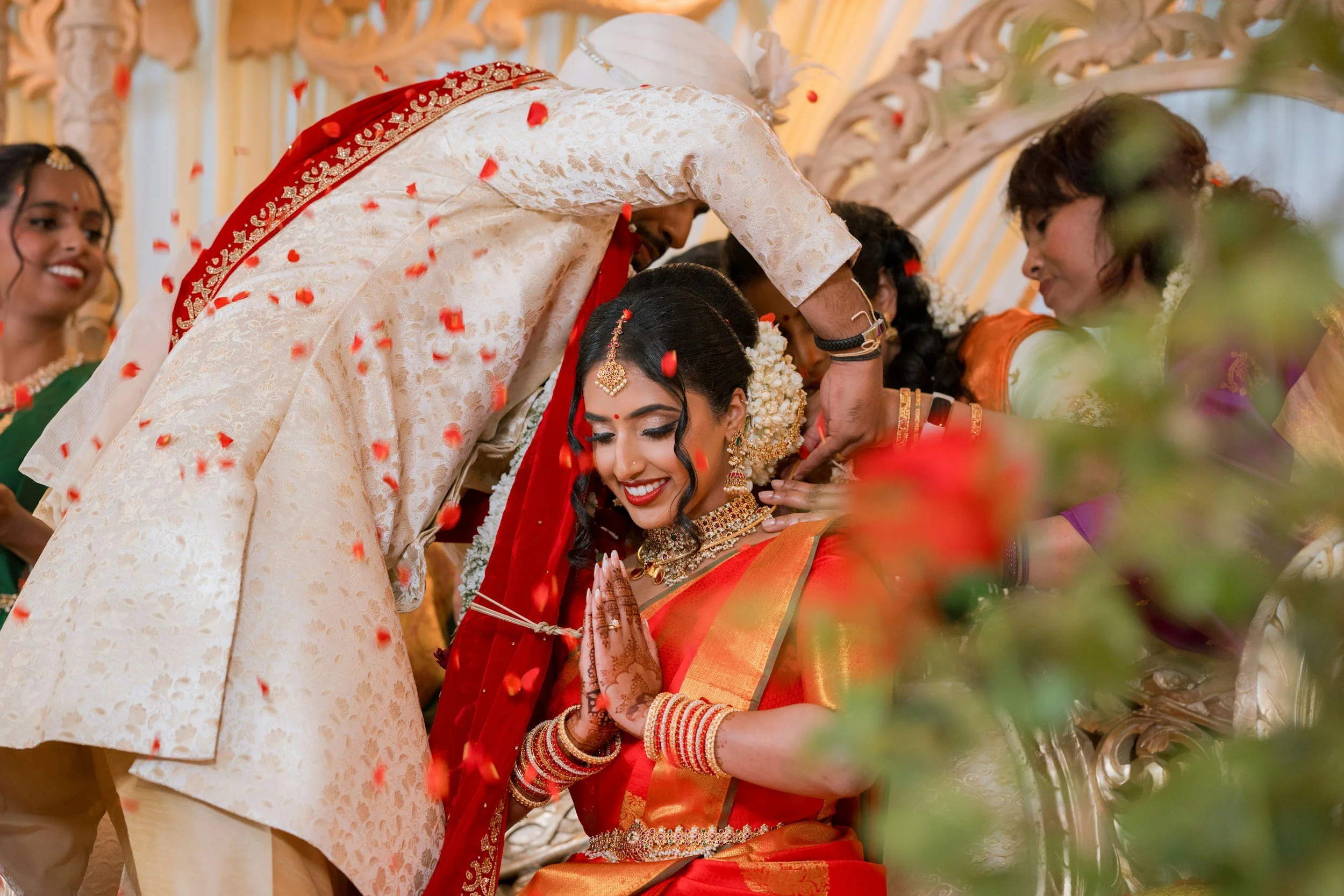 Indian wedding ceremony with bride and groom in traditional attire, surrounded by women in colorful sarees, flowers, and rose petals.