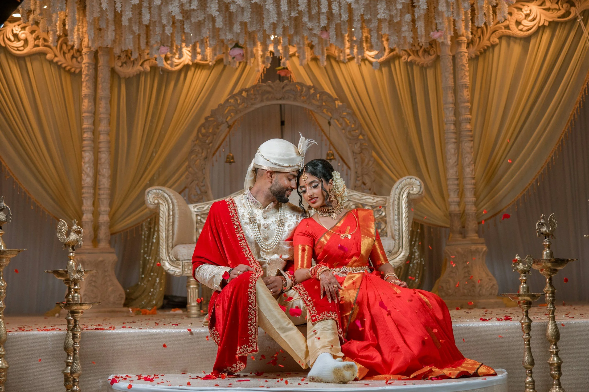 A newly married Indian couple dressed in traditional wedding attire sitting closely on a decorated stage, surrounded by floral decorations and ornate draping.