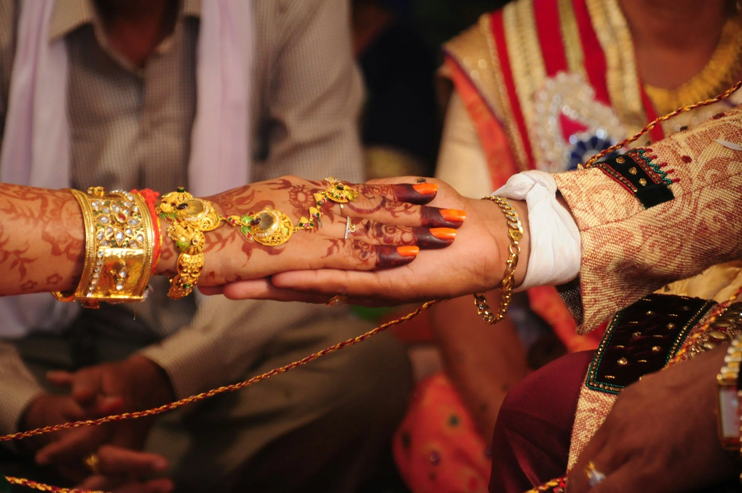 Close-up of hands covered in henna, wearing gold jewelry, holding hands during a traditional Indian wedding ceremony. In the background, people dressed in traditional attire.