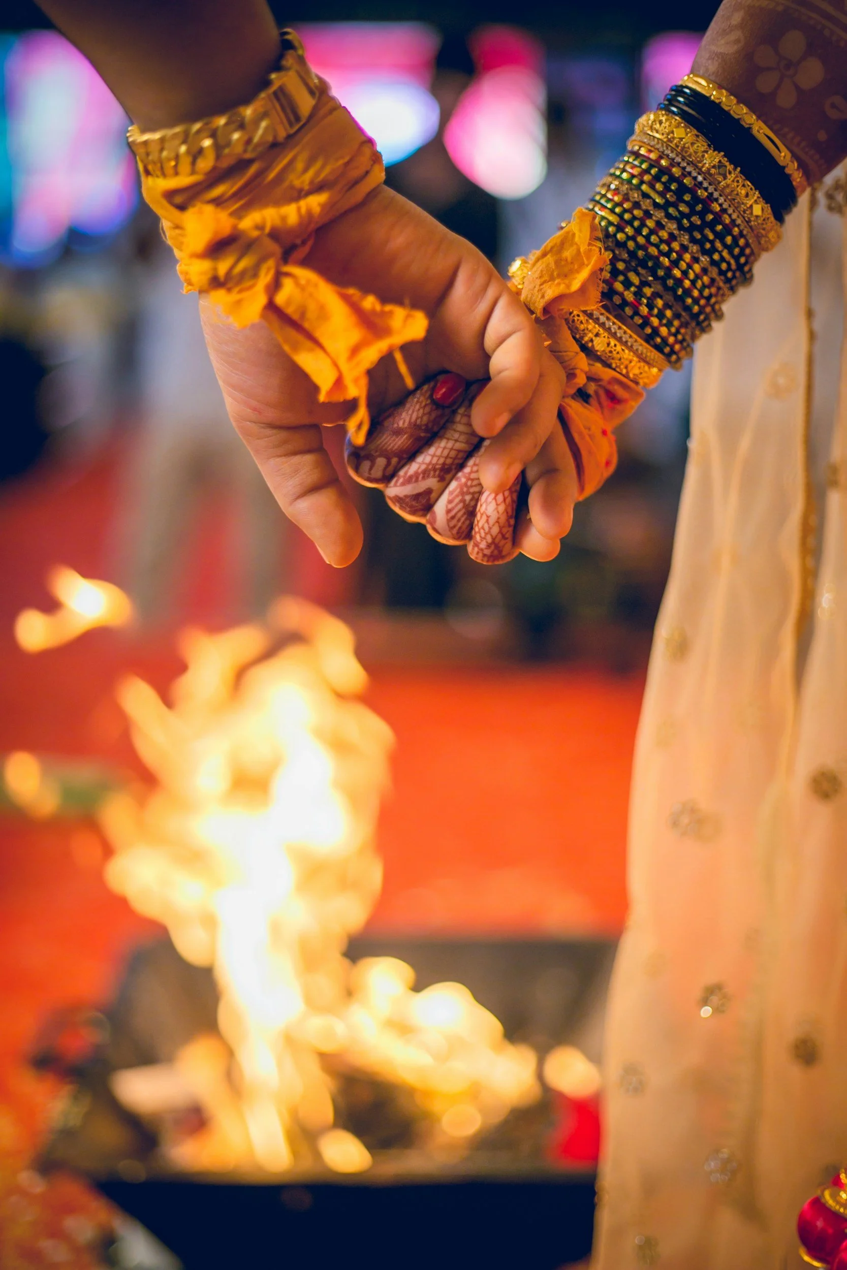 Hindu wedding ritual with two hands joined, one decorated with orange cloth and multiple colorful bangles. Fire in front indicates a traditional ceremony.