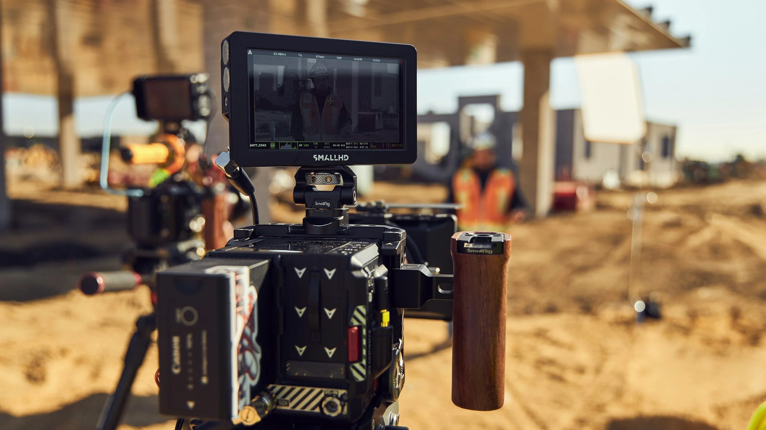 Close-up of a professional video camera filming a construction site, with blurred workers and structures in the background.