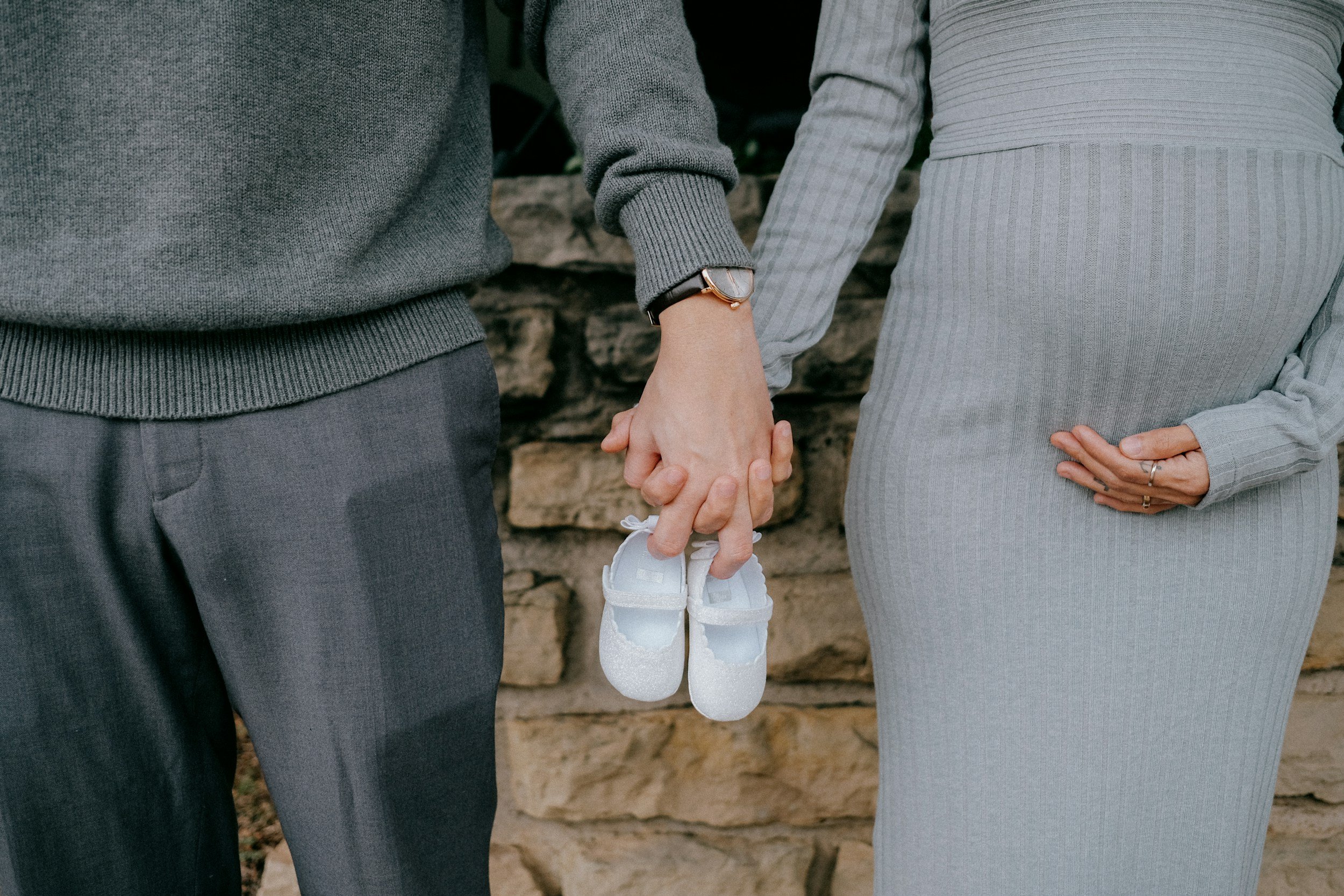 A couple holding hands with small baby shoes hanging from one hand, dressed in gray and brown clothing, outdoors near a stone wall.