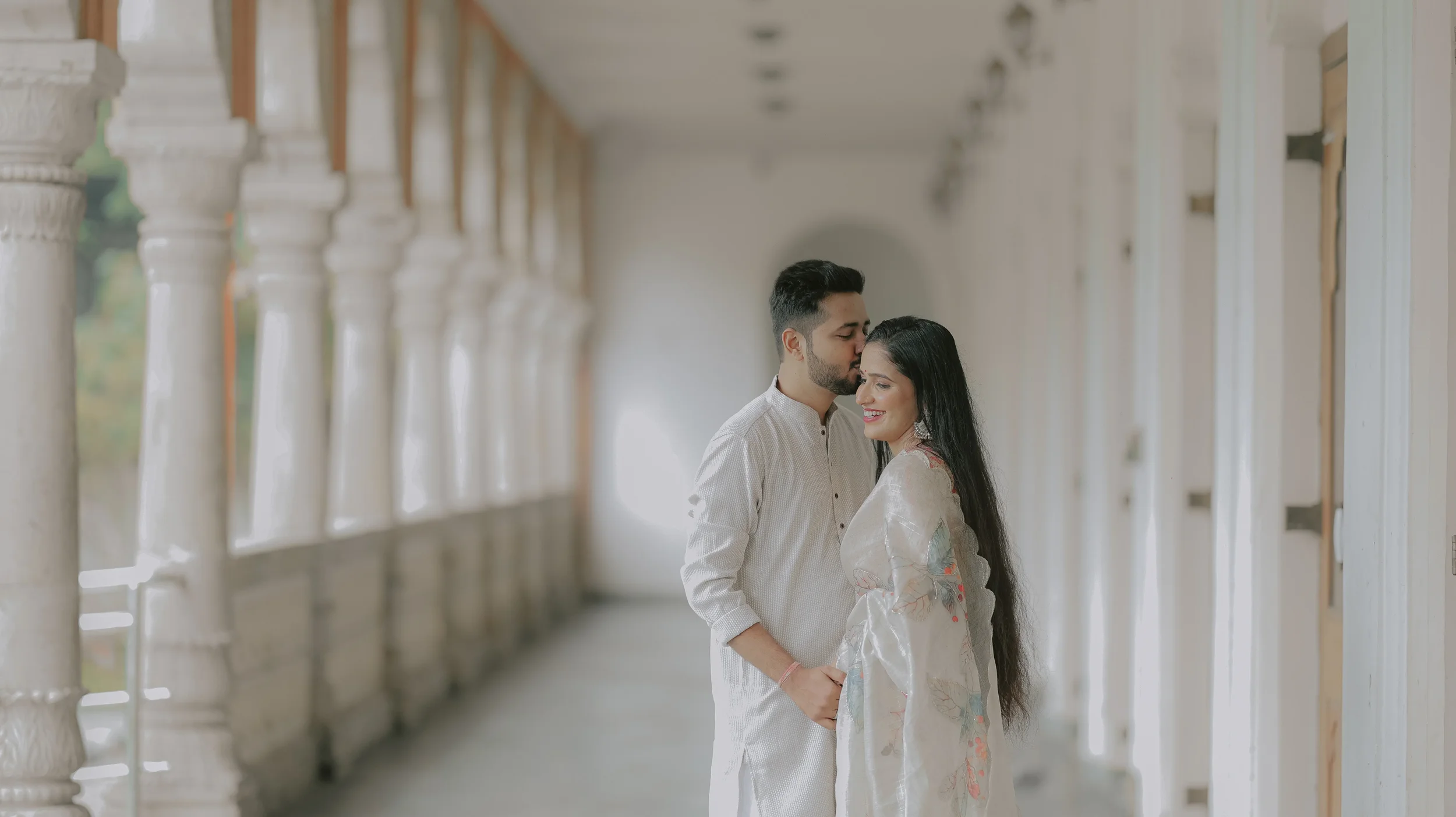 A couple standing close together in a corridor with white columns, smiling and sharing a tender moment.
